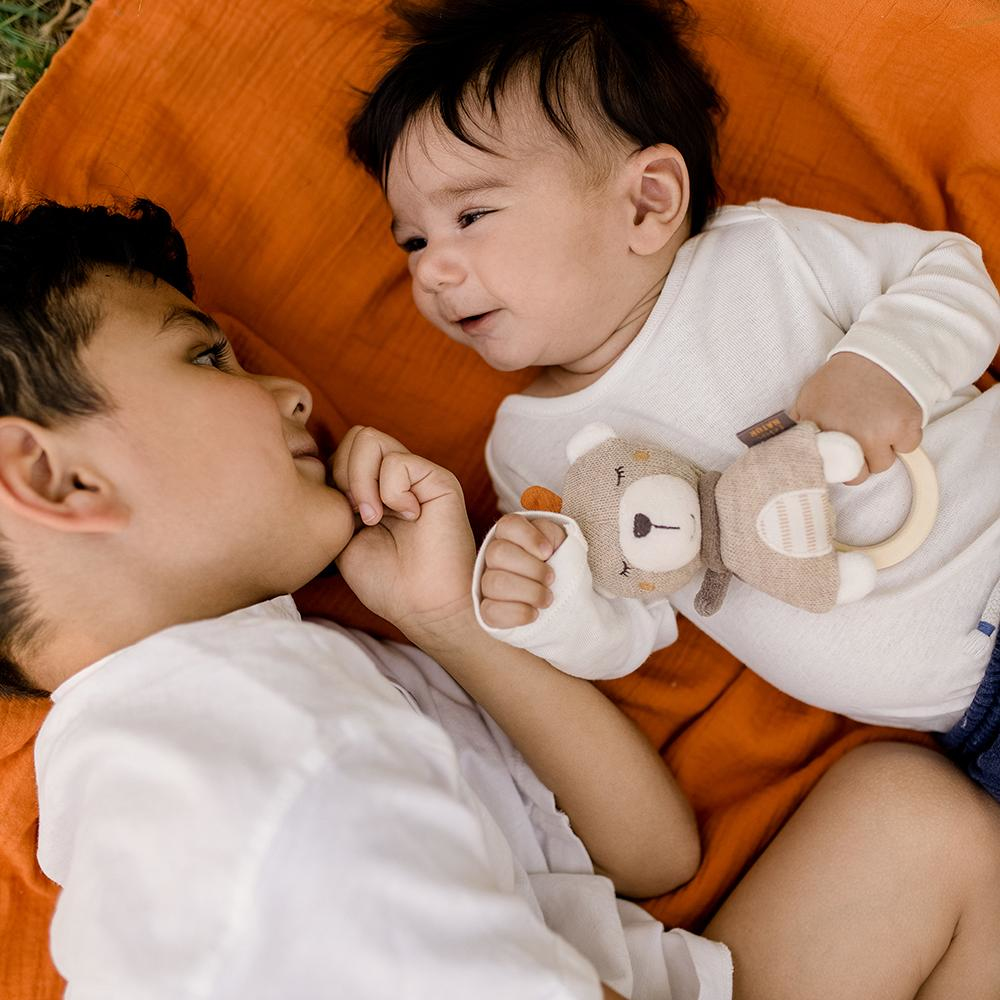 Due bambini sdraiati su una coperta arancione, uno con un orsacchiotto, si guardano.