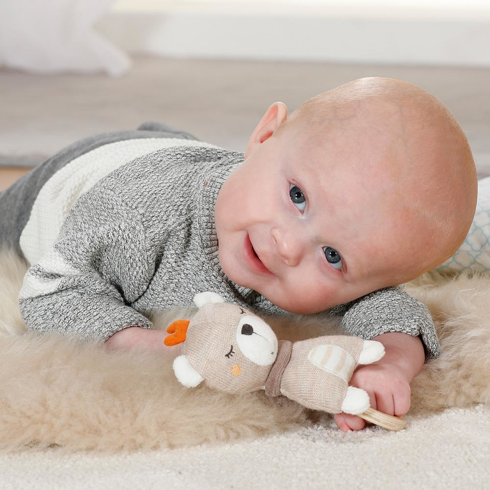 Bambino sdraiato su una superficie simile alla pelliccia, guarda un orsacchiotto di peluche, con sfondo neutro.