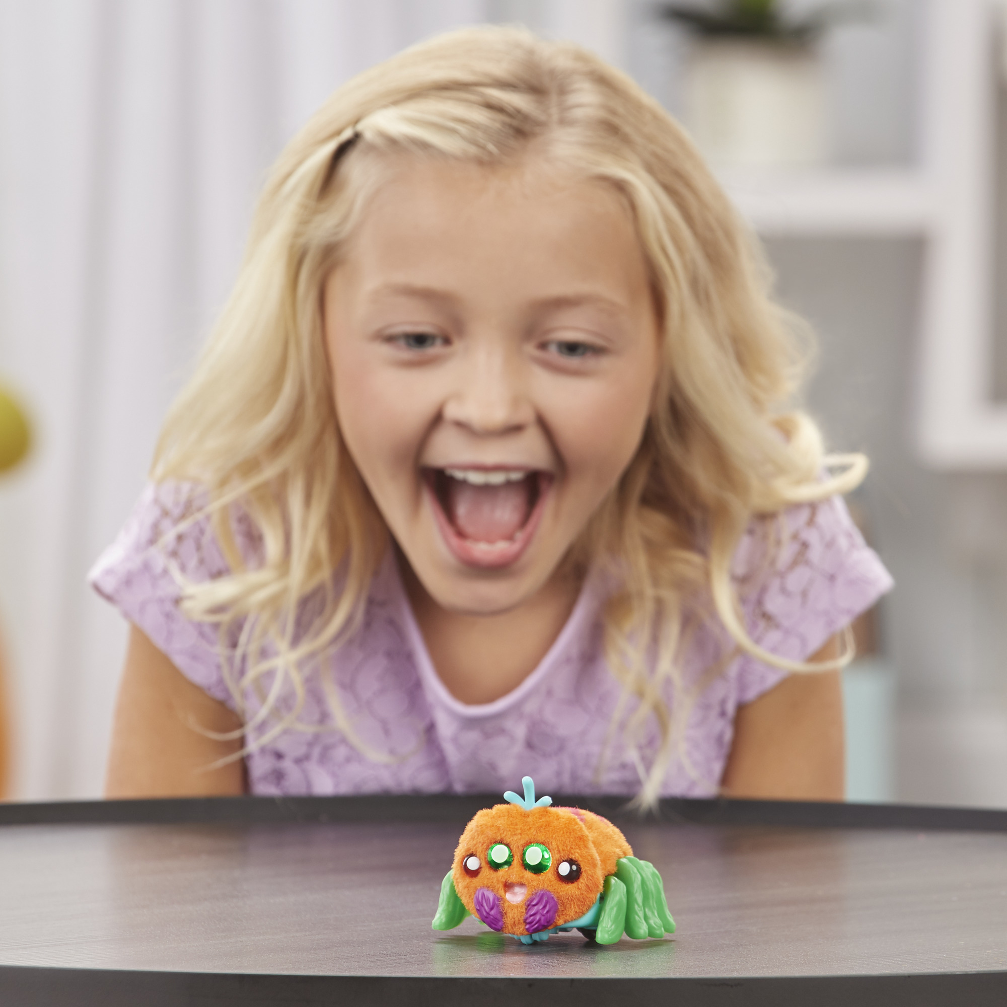 Blondes Mädchen mit offenem Mund beobachtet Spielzeugspinne auf einem Tisch. Hintergrund unscharf.