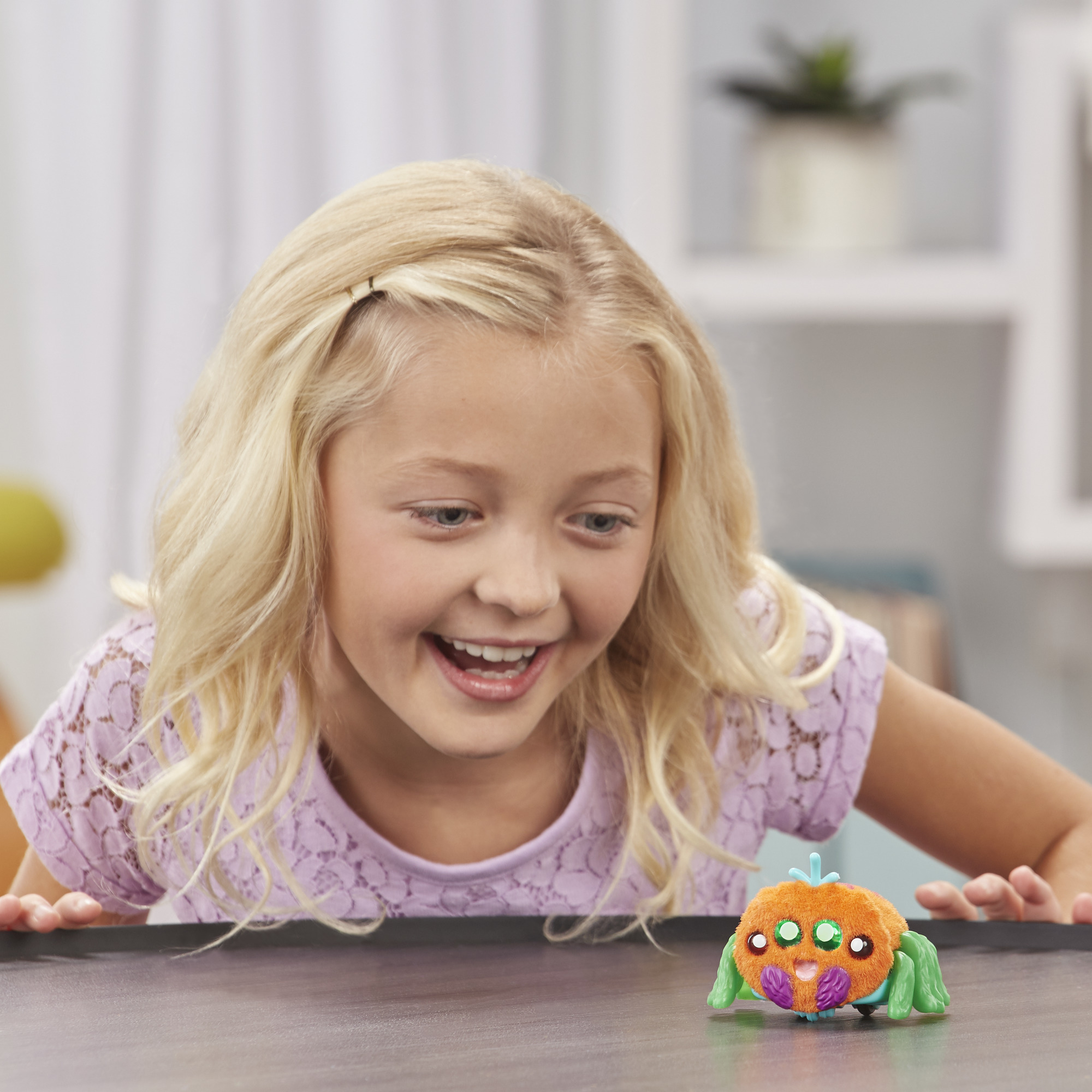 Blondes Mädchen lächelt einen Spielzeug-Spinne an. Orange mit grünen Beinen.