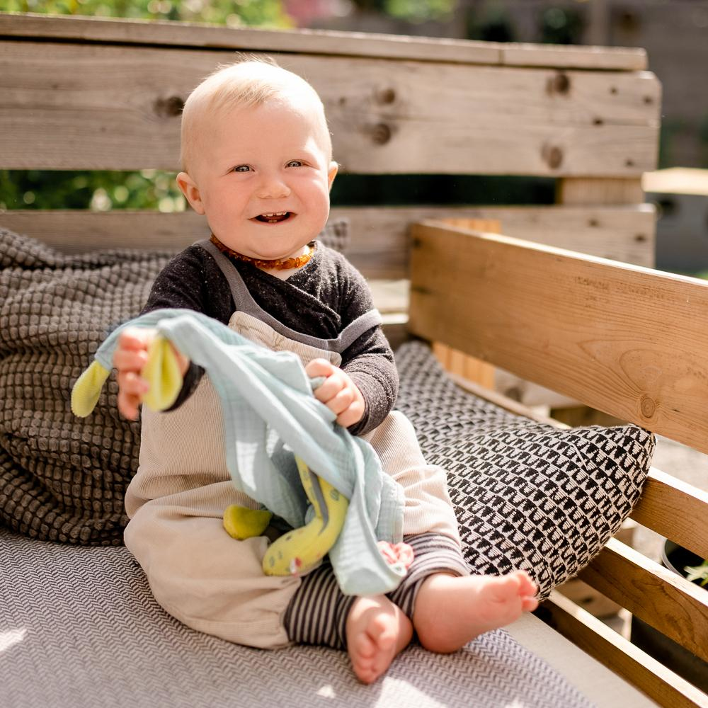 Niemowlę siedzi na ławce trzymając pluszową zabawkę. Niemowlę jest blondynem. Ustawienie na zewnątrz.