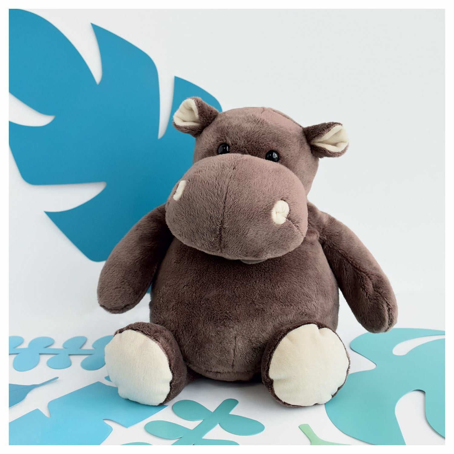 Peluche ippopotamo marrone con dettagli bianchi seduto davanti a foglie.