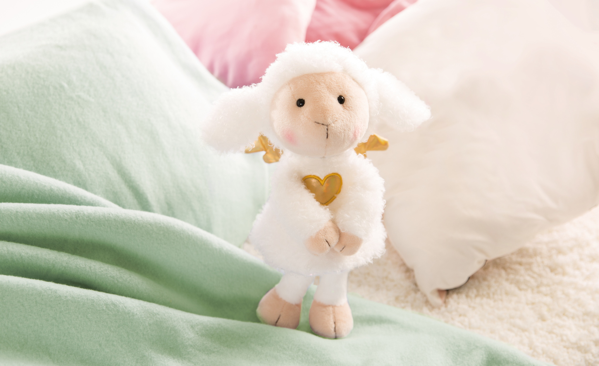 Biała pluszowa owca ze złotymi skrzydłami i sercem. Leżąca na jasnozielonym kocu i poduszkach.