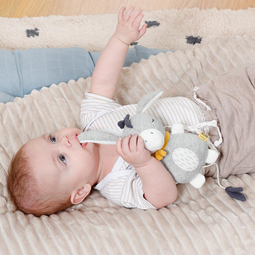 Ein Baby liegt auf einem Bett und spielt mit einem grauen Plüschtier. Beiger Hintergrund.