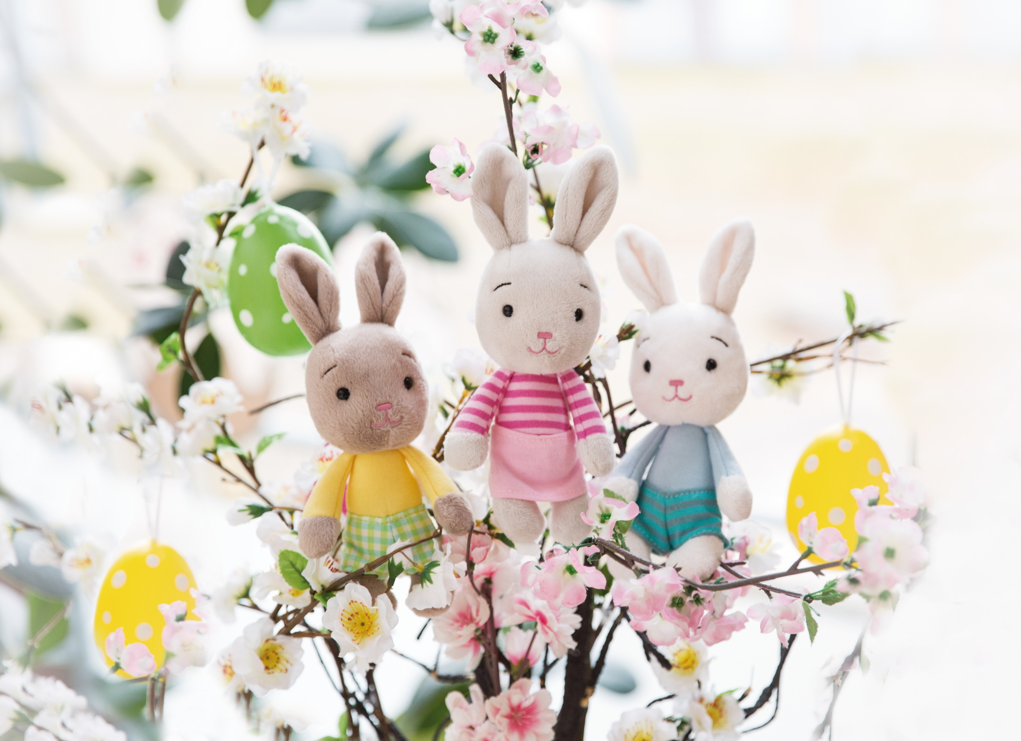 Trzy pluszowe króliczki siedzące na gałęziach z kwiatami i pisankami. Jasne tło.