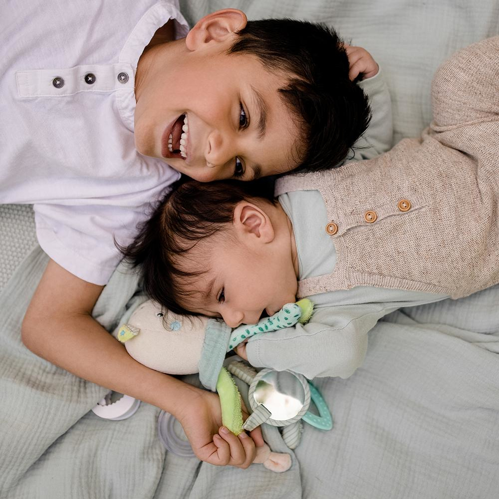 Dwoje dzieci, jedno przytulające pluszową zabawkę, leżą razem na kocu, uśmiechając się.