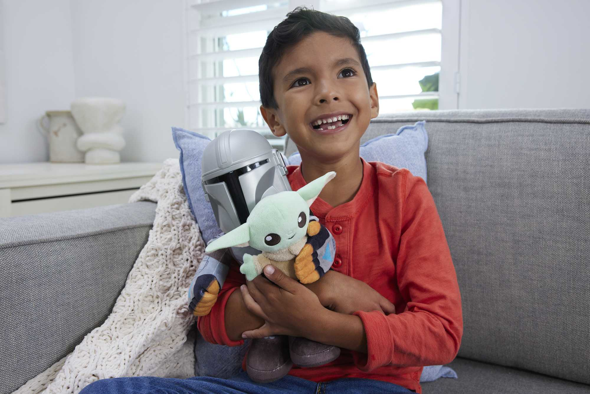 Chłopiec uśmiecha się, trzymając pluszaki Mandalorian i Grogu, siedząc na kanapie.