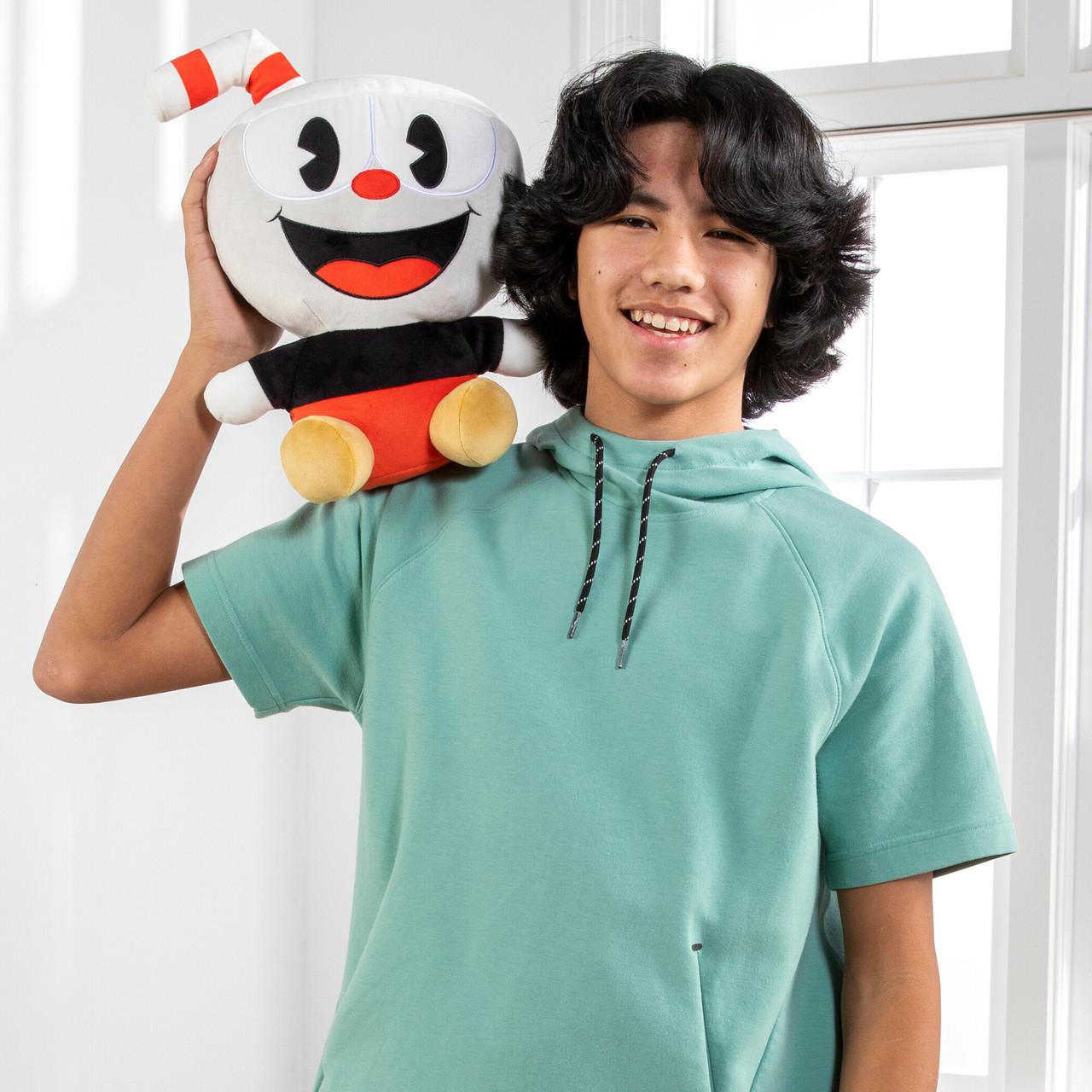 Un adolescente tiene un peluche di Cuphead. Sorride, indossa una felpa con cappuccio verde.