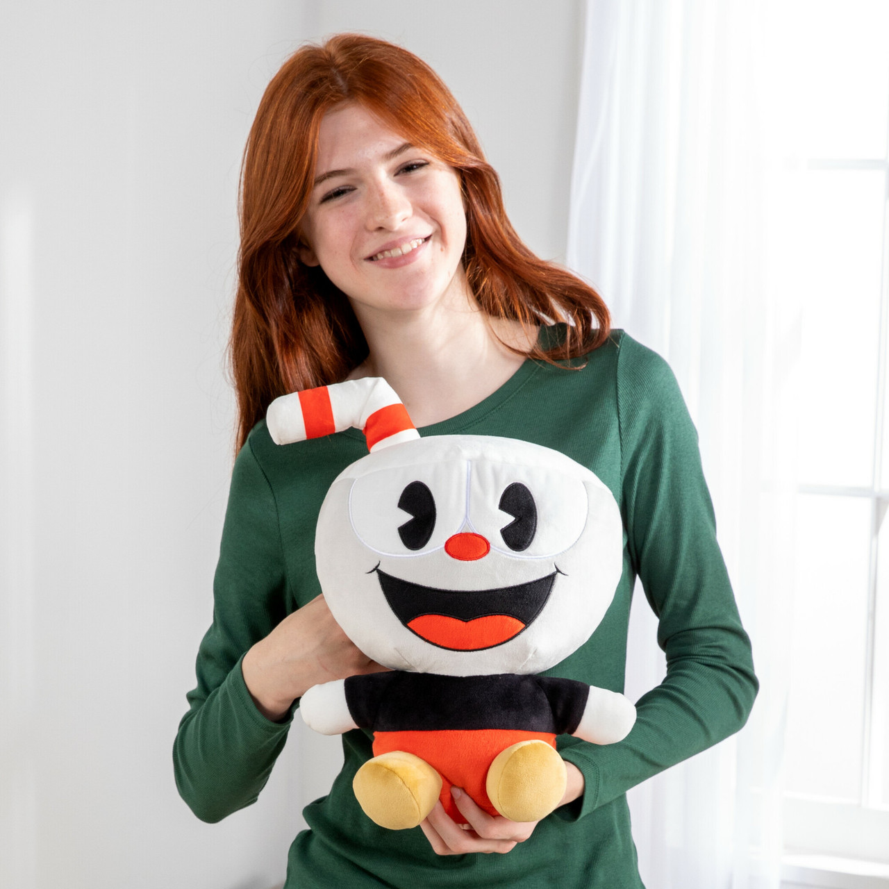Una donna tiene un peluche di Cuphead. Sorride, indossa una camicia verde a maniche lunghe.