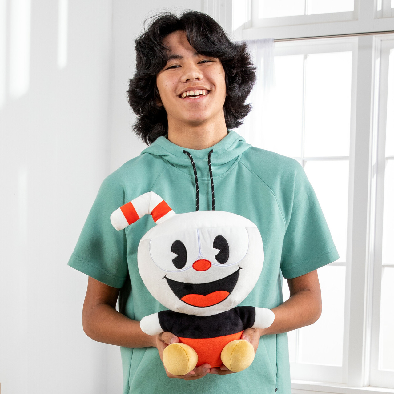 Un adolescente tiene un peluche di Cuphead. Sorride, con una felpa verde, vicino a una finestra.