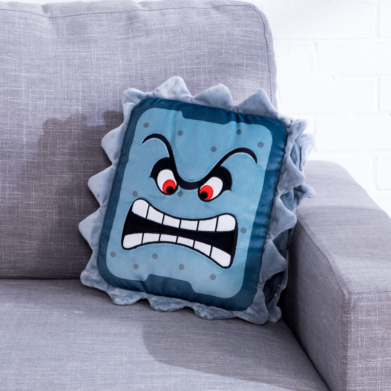Coussin Thwomp au visage en colère sur un canapé gris, fond de mur en briques blanches.