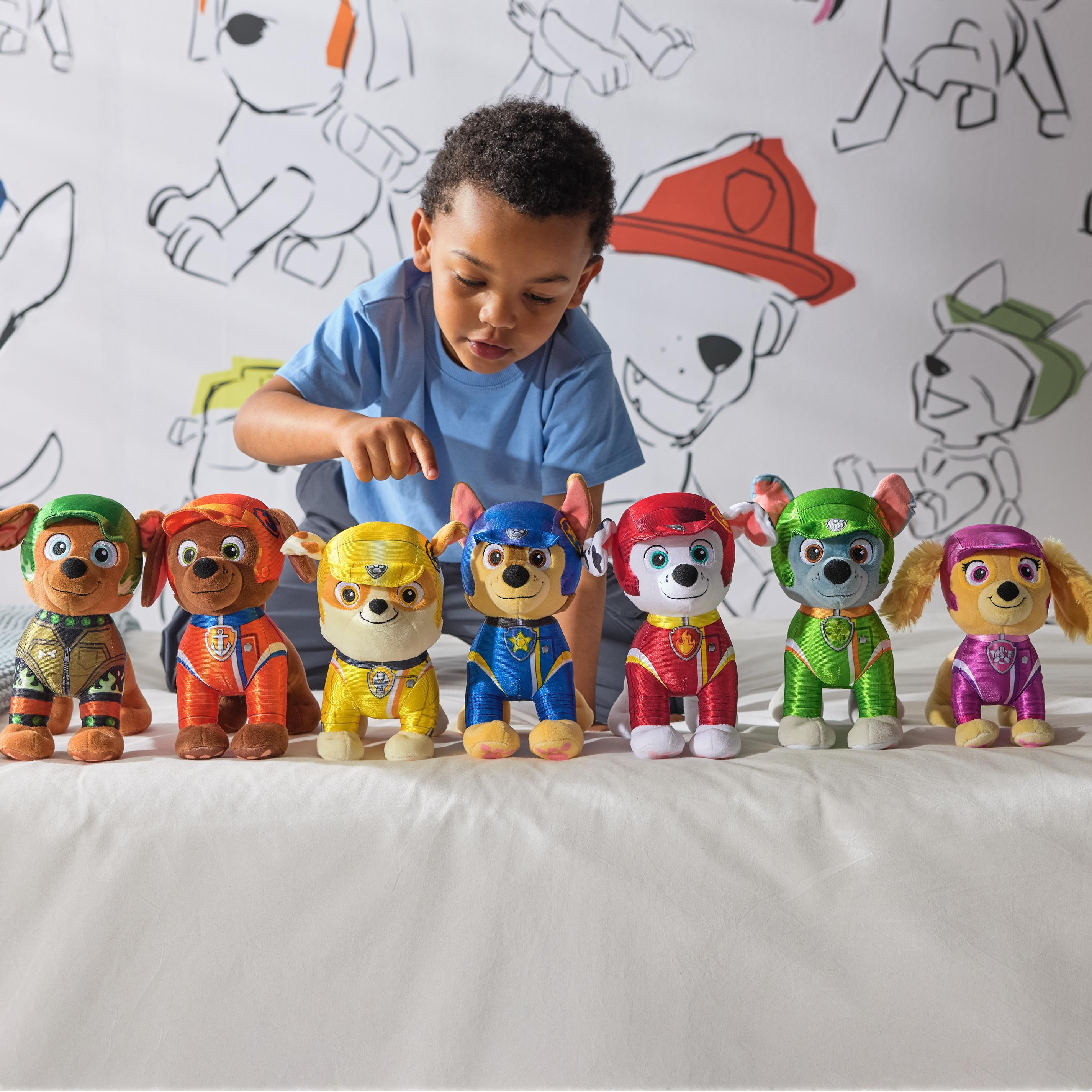 Ein Kind zeigt auf sieben Paw Patrol Plüschtiere auf weißer Oberfläche, mit thematischem Hintergrund.