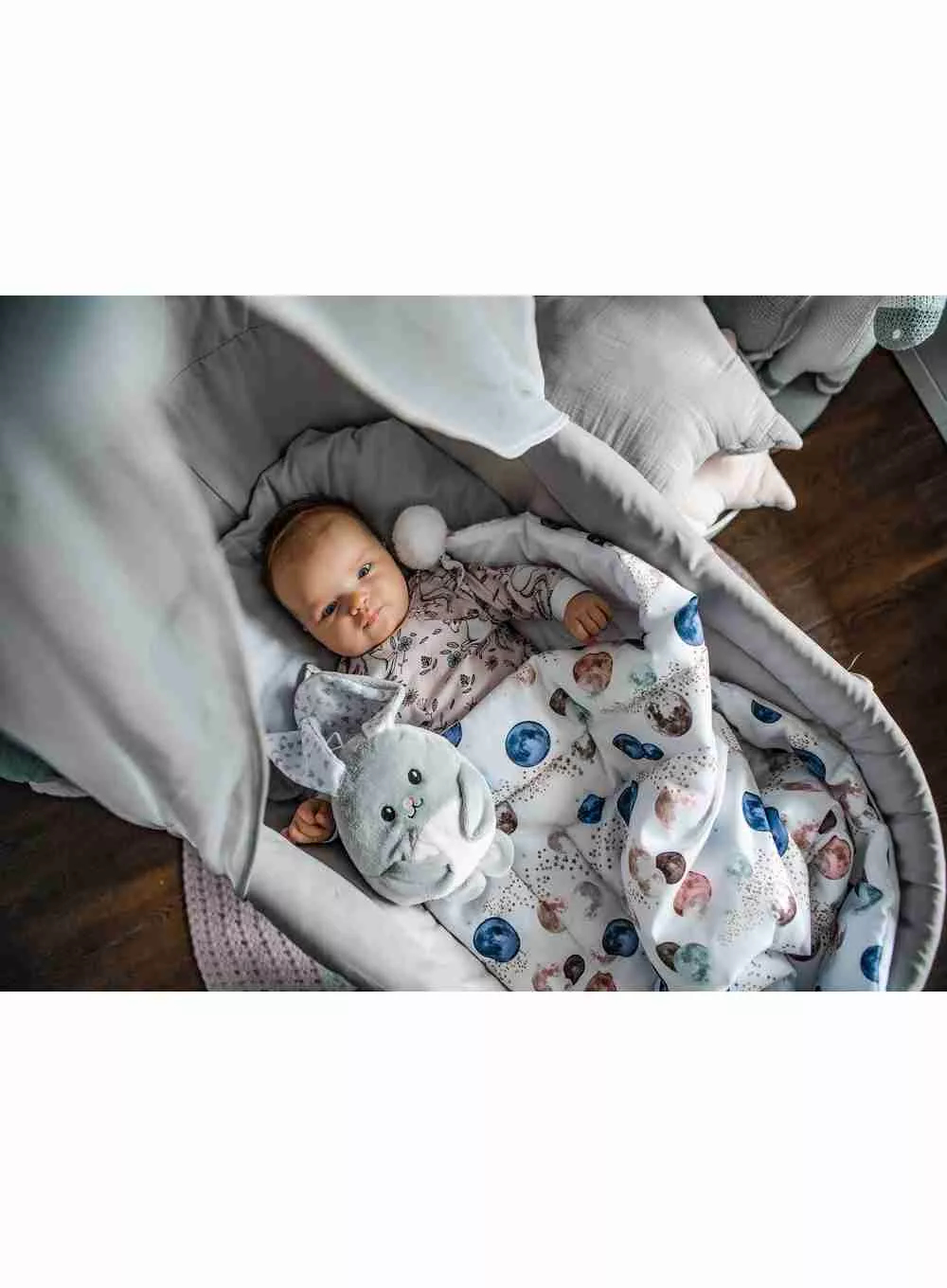 Un bambino in una culla grigia, coperto da una coperta, con un coniglietto di peluche.