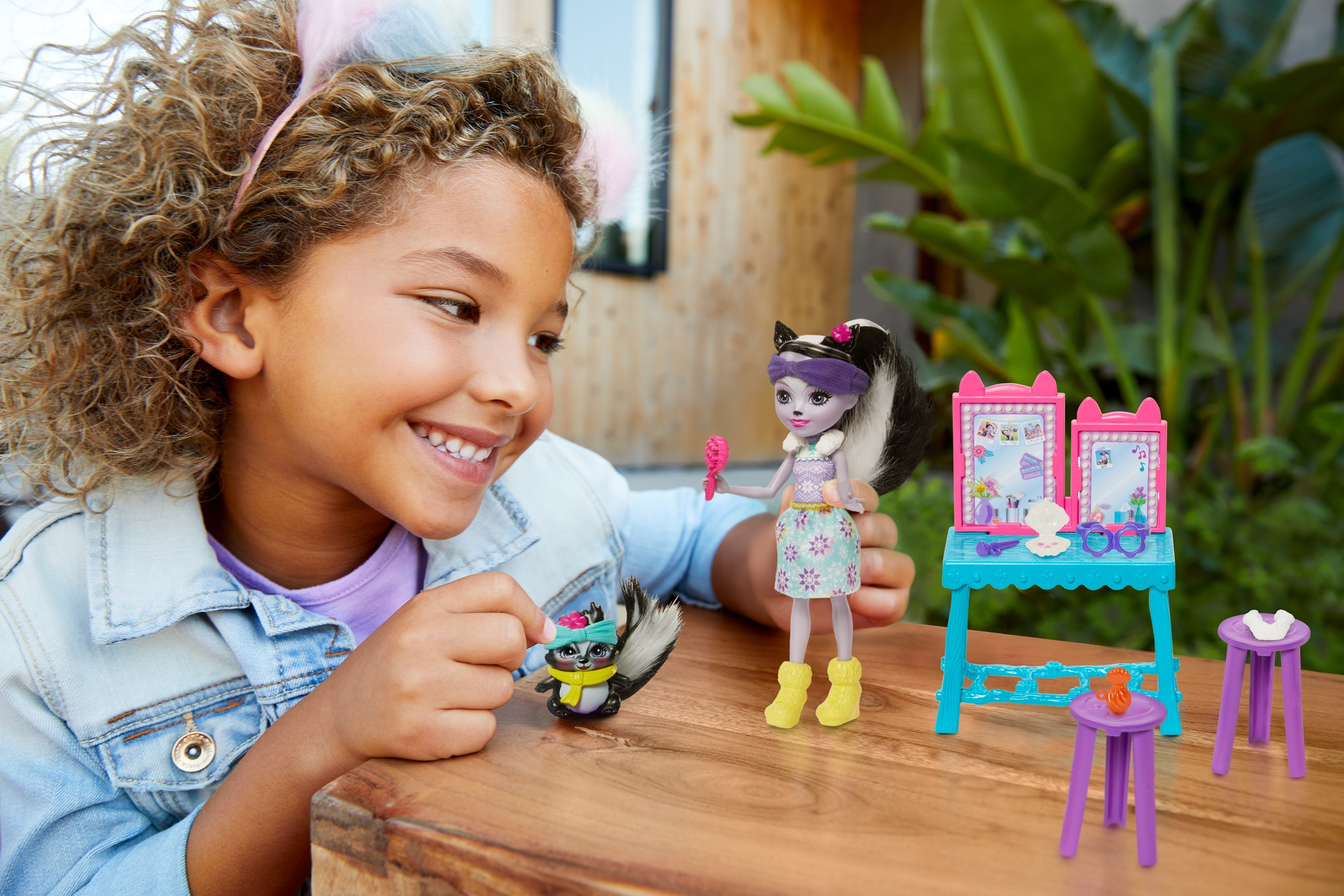 Ein Mädchen lächelt und hält eine kleine Puppe und ein Stinktier-Spielzeug. Andere Spielzeuge und ein Tisch sind in der Szene.