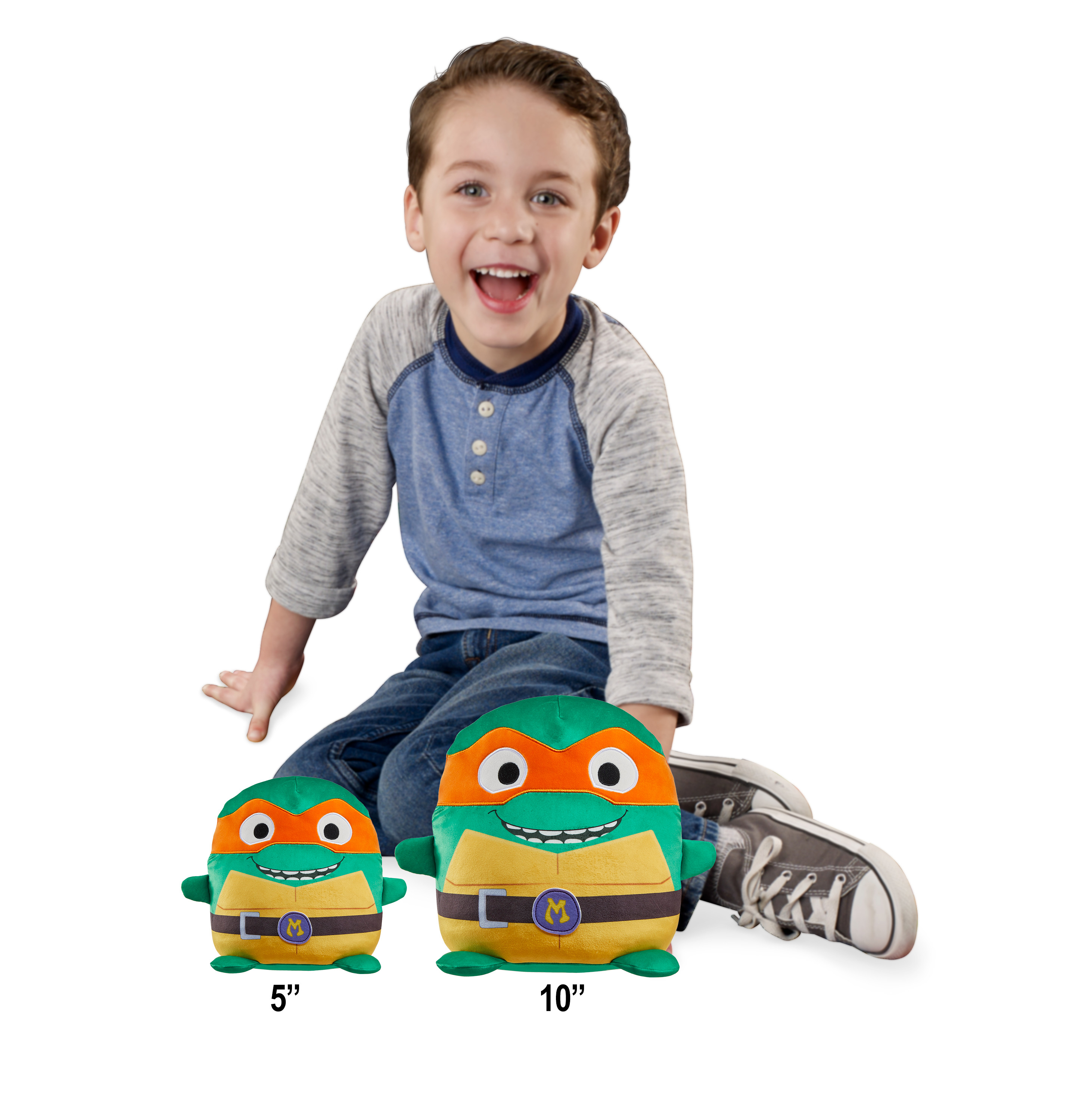 Un bambino sorridente, seduto accanto a due peluche Tartarughe Ninja di diverse dimensioni.
