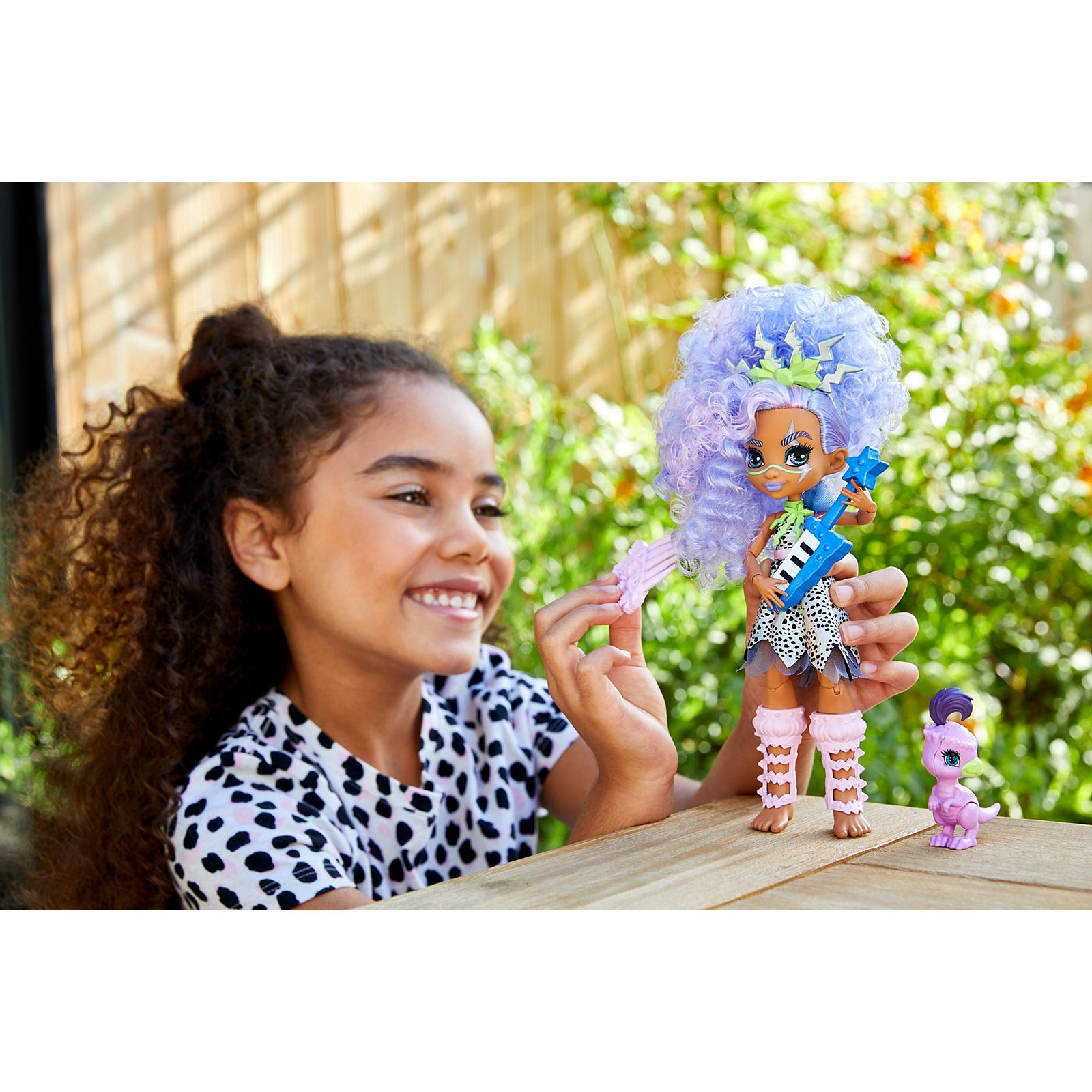 Dziewczyna uśmiecha się, bawiąc się lalką trzymającą gitarę. W pobliżu siedzi mały fioletowy dinozaur.