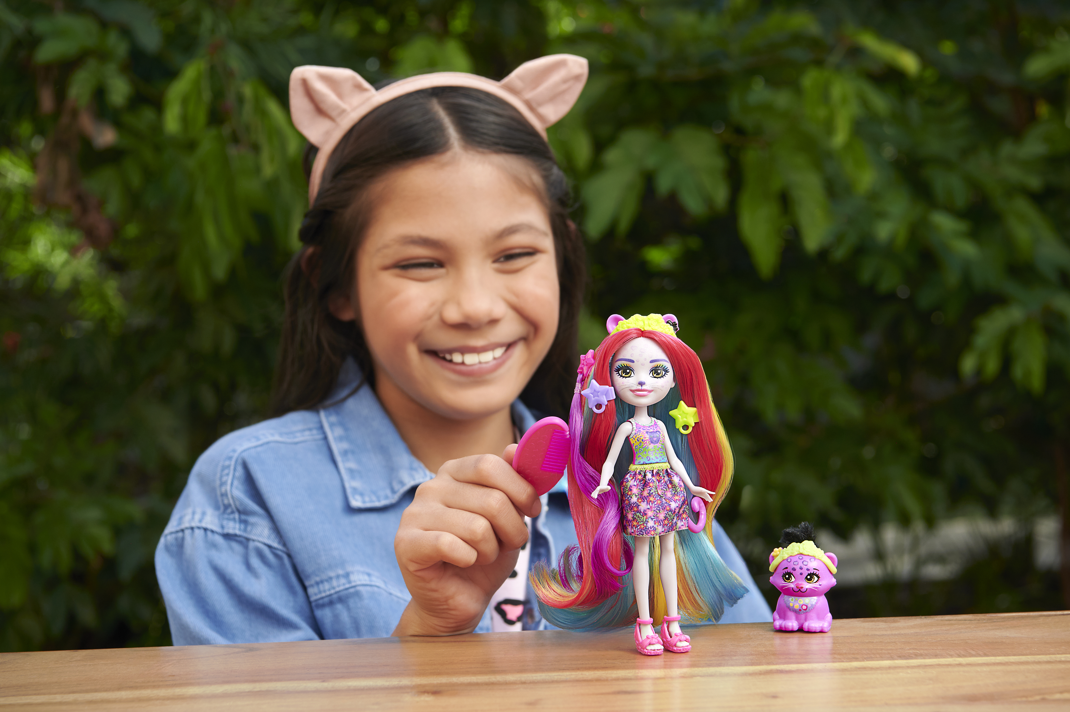 Dziewczyna uśmiecha się do lalki. Lalka ma kocie uszy i kolorowe włosy. Obok niej zabawkowy miś.