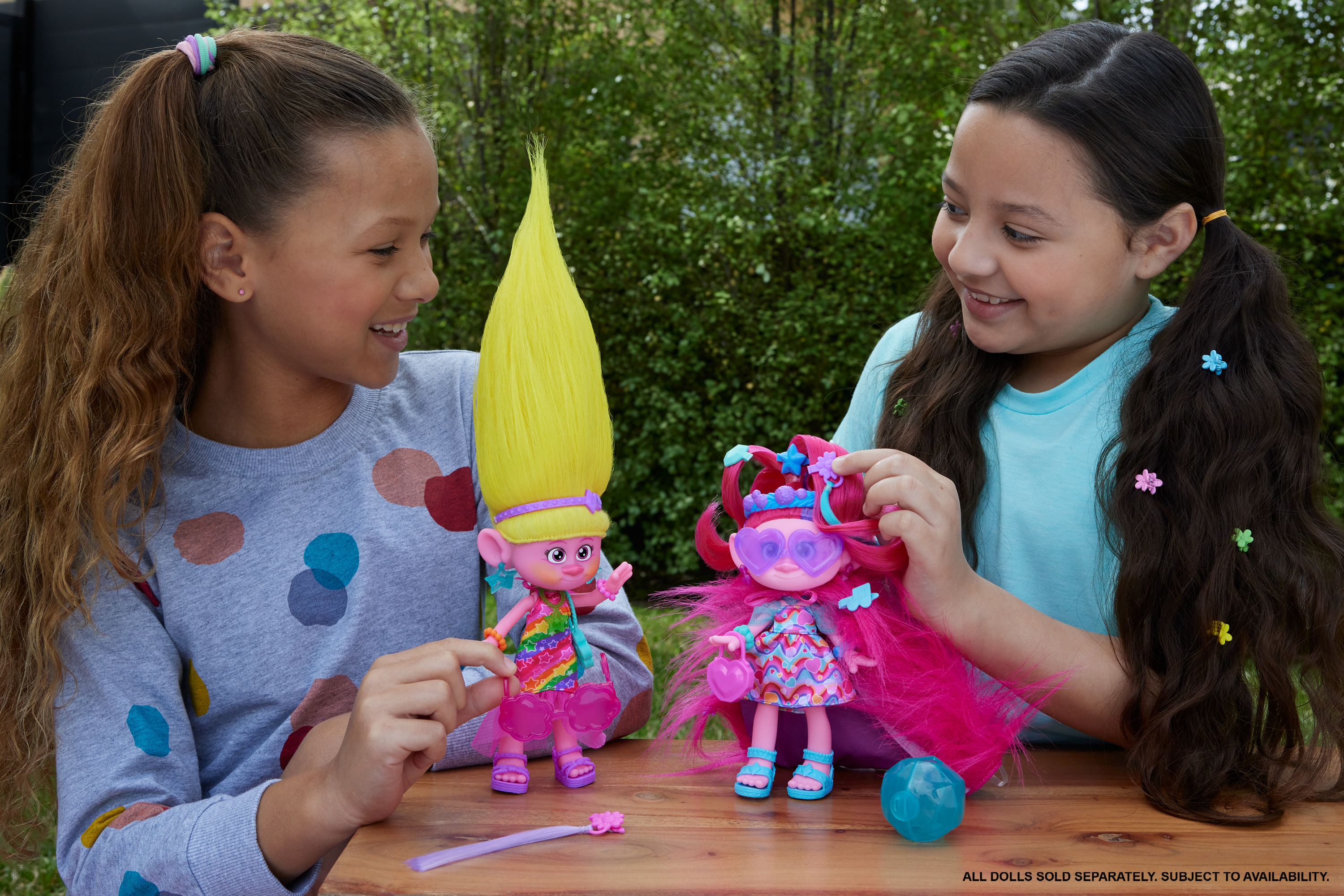 Due ragazze giocano con bambole Troll. Una bambola ha i capelli gialli, l'altra ha i capelli rosa. Sono presenti vari accessori.