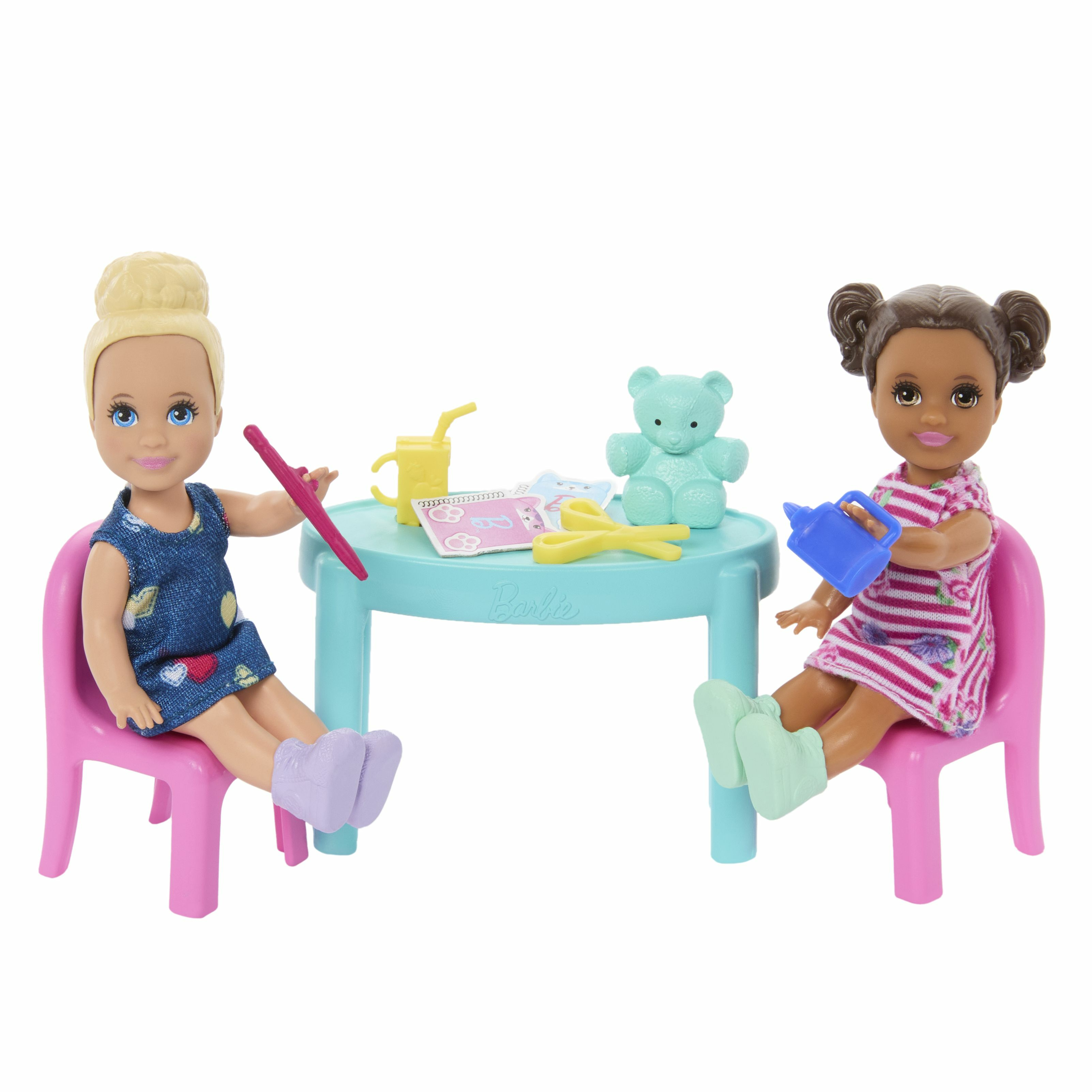 Dos muñecas sentadas en una mesa con juguetes, en un fondo blanco.