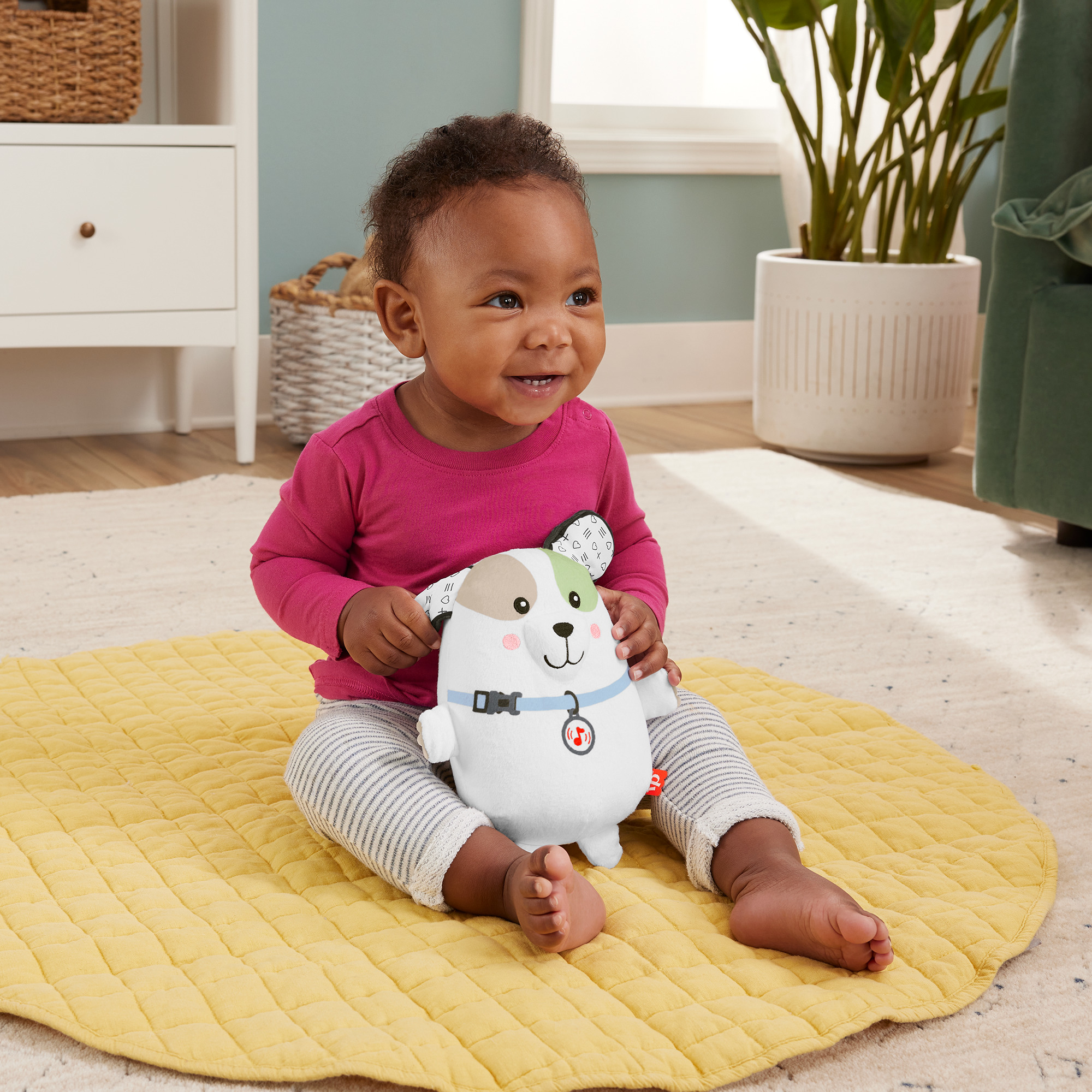 Ein Baby sitzt auf einer gelben Matte und hält ein Plüschhundespielzeug. Das Baby lächelt.