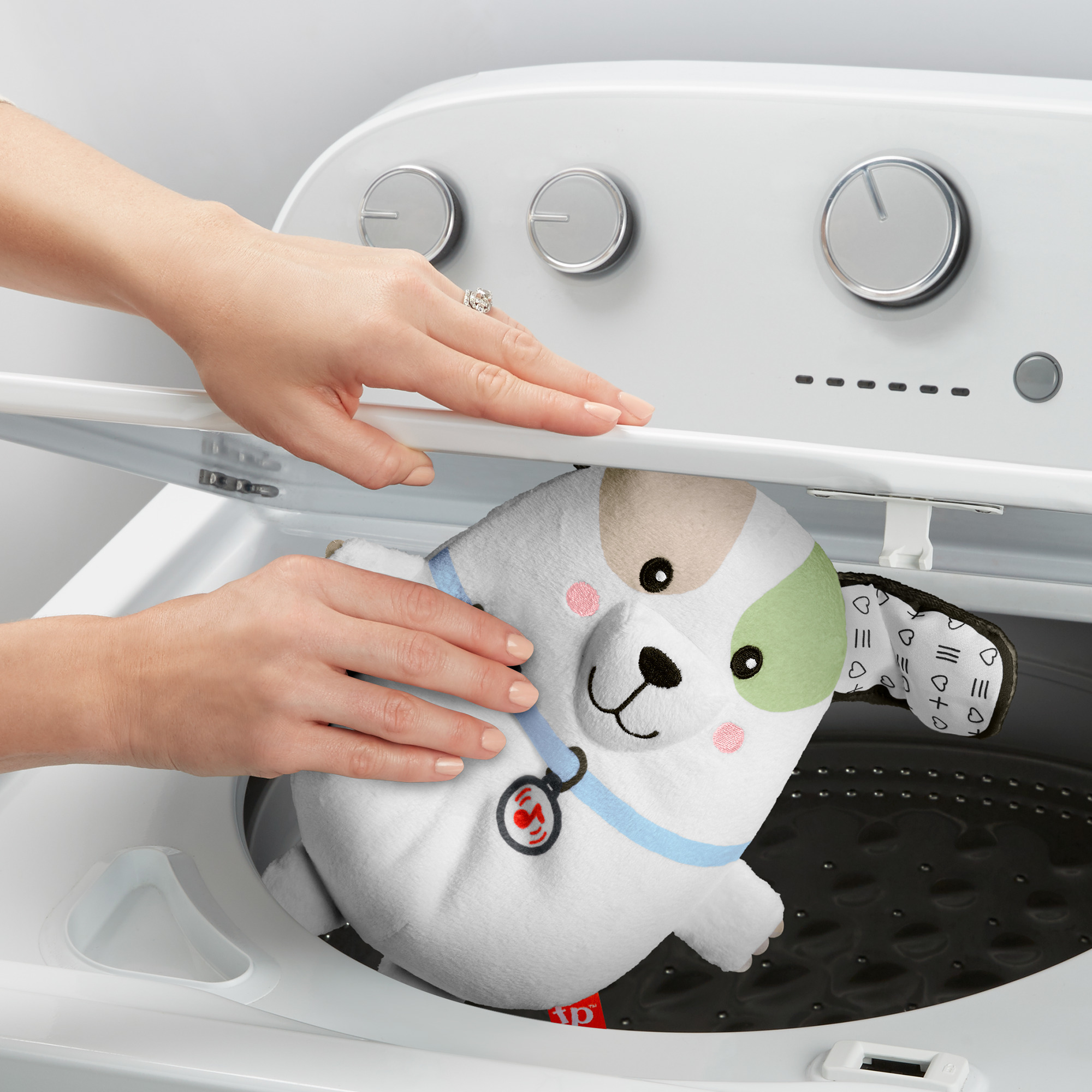 Hände, die ein Hundekuscheltier in eine Waschmaschine legen. Das Spielzeug ist weiß, mit grünen und schwarzen Akzenten.