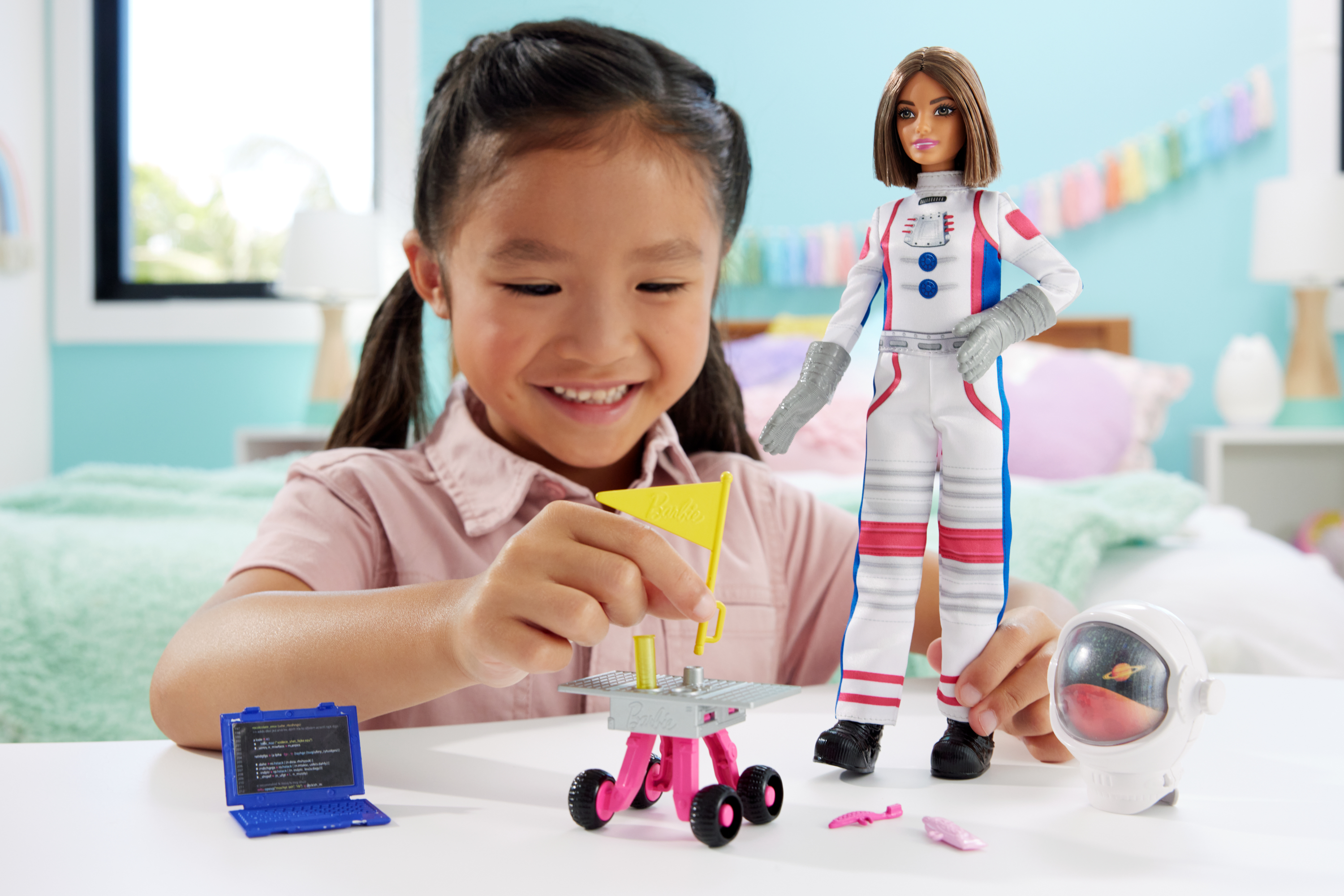 Mała dziewczynka bawiąca się lalką Barbie astronautką i akcesoriami o tematyce kosmicznej. W pobliżu laptop.