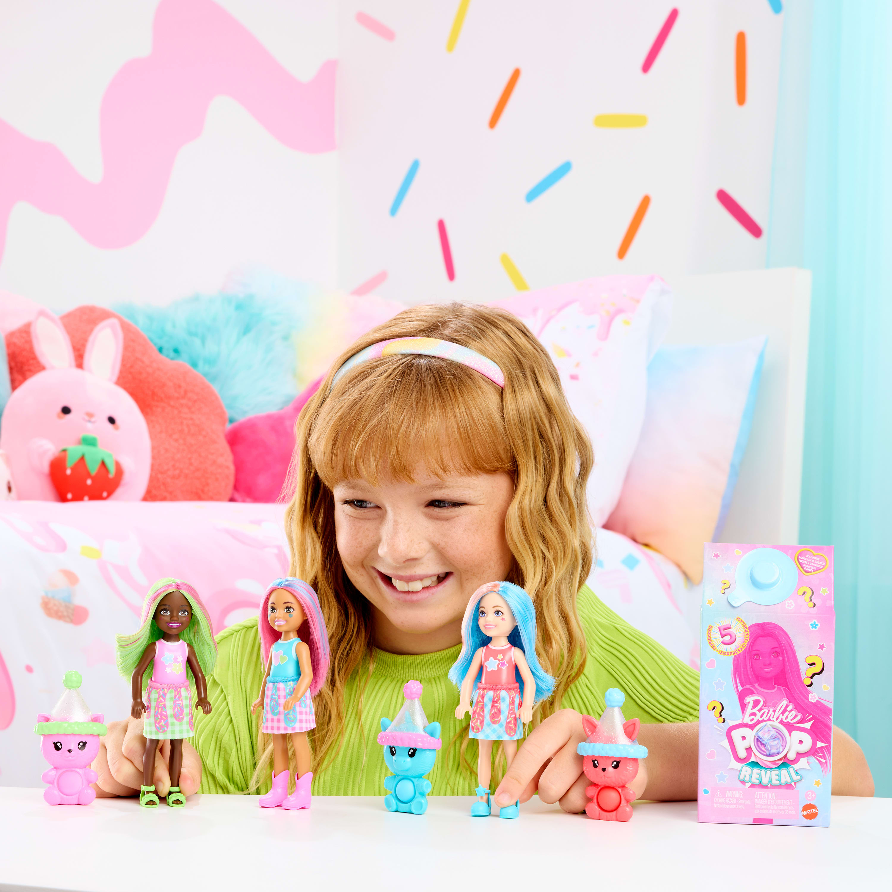 Mädchen lächelt Barbie Pop Reveal-Puppen an. Puppen haben bunte Haare und Outfits. Zubehör liegt auf dem Tisch.