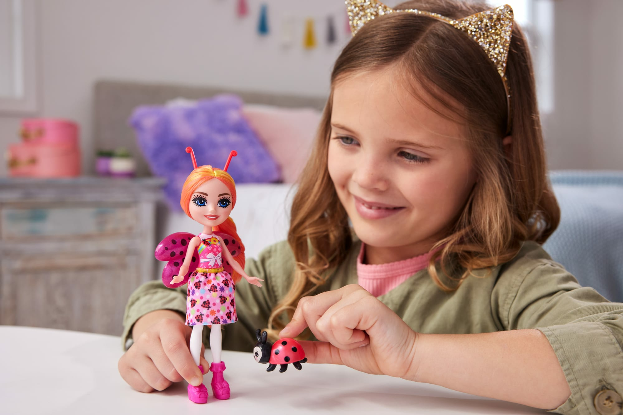 Ein junges Mädchen mit einer Puppe in der Hand, neben einem Marienkäfer-Spielzeug, lächelt breit beim Spielen.