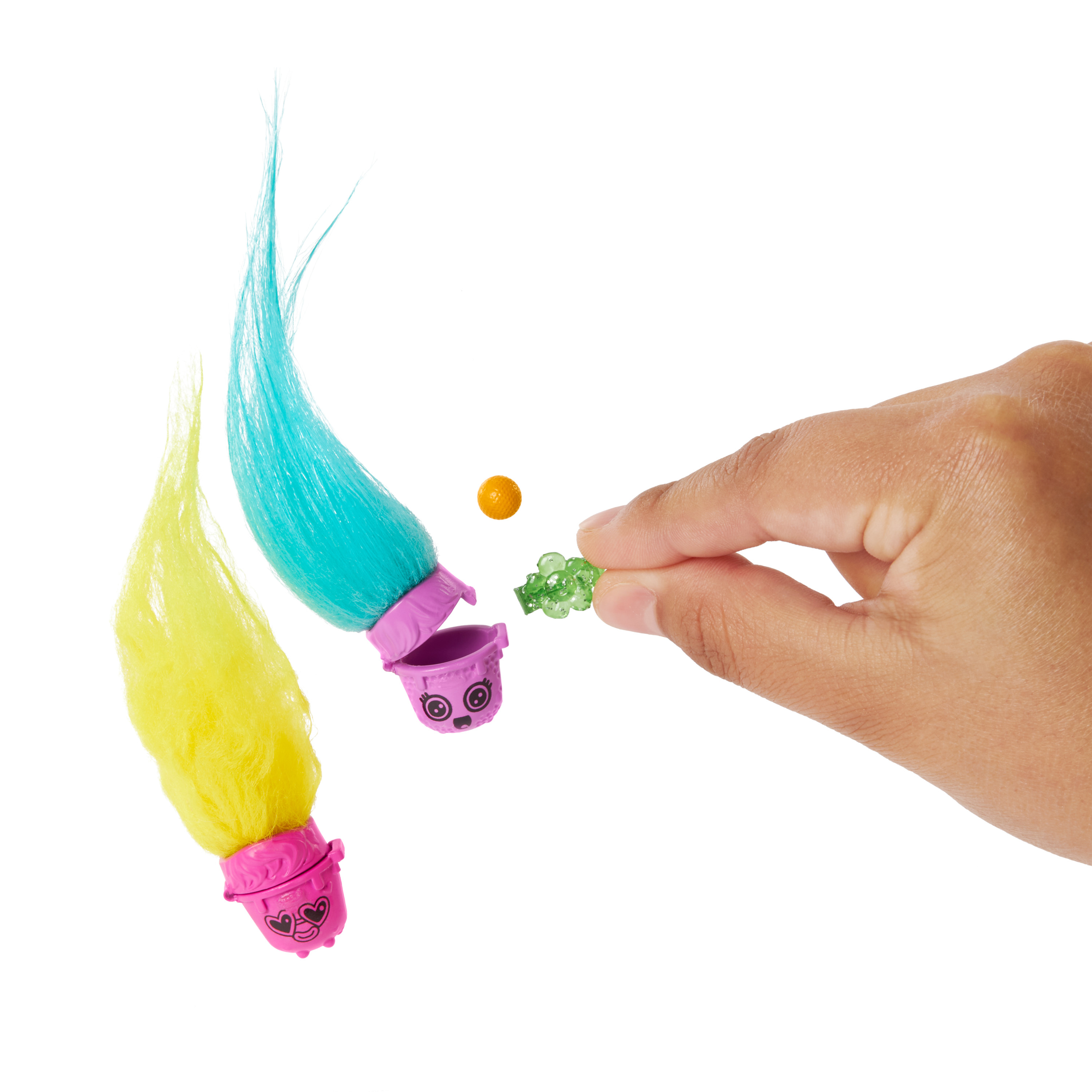 Ręka trzymająca mały zielony przedmiot. Dwie lalki trolli z kolorowymi włosami i małymi pojemnikami. Białe tło.