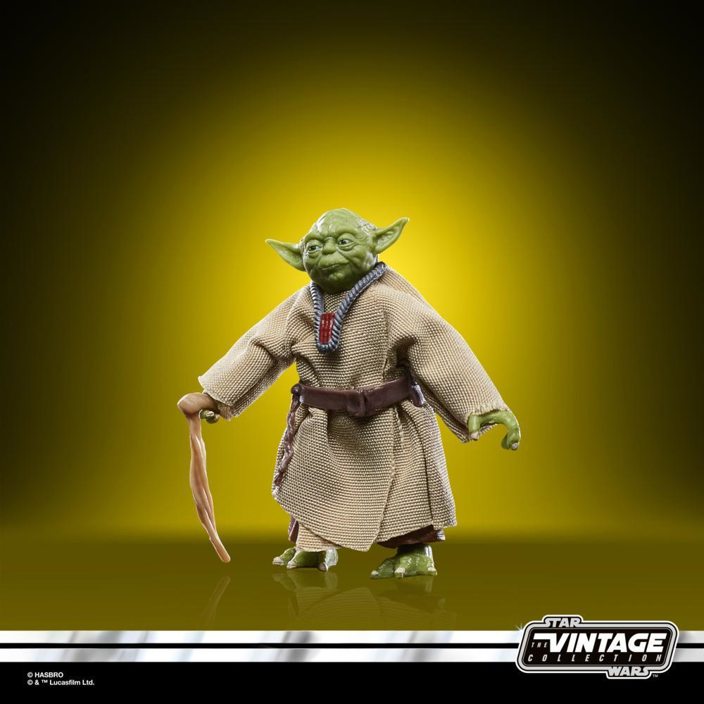 Yoda figürü, yeşil ten, kahverengi cübbe, baston tutuyor, sarı bir arka plan üzerinde duruyor.