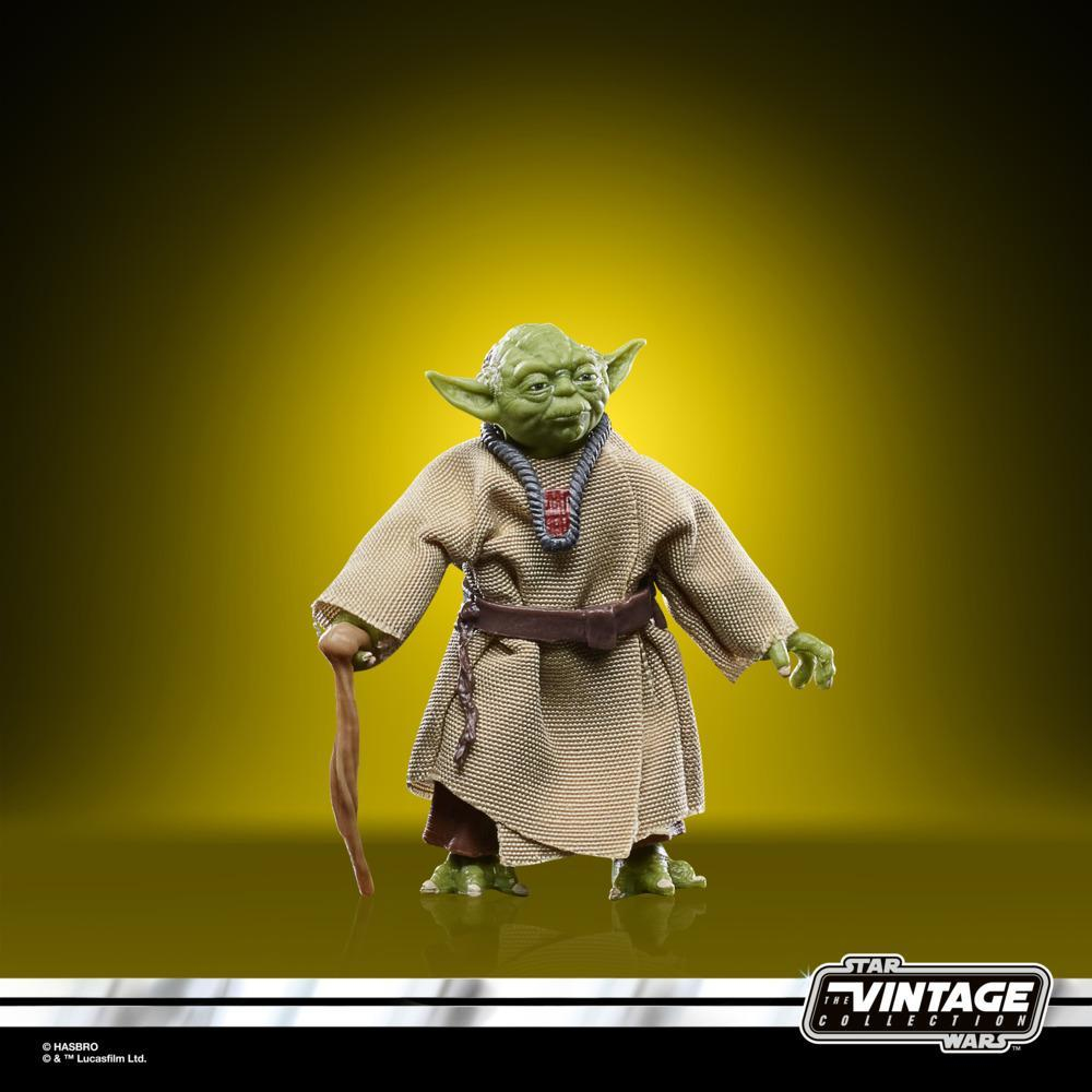 Yoda figürü, yeşil ten, kahverengi cübbe, baston tutuyor, sarı bir arka plan üzerinde duruyor.