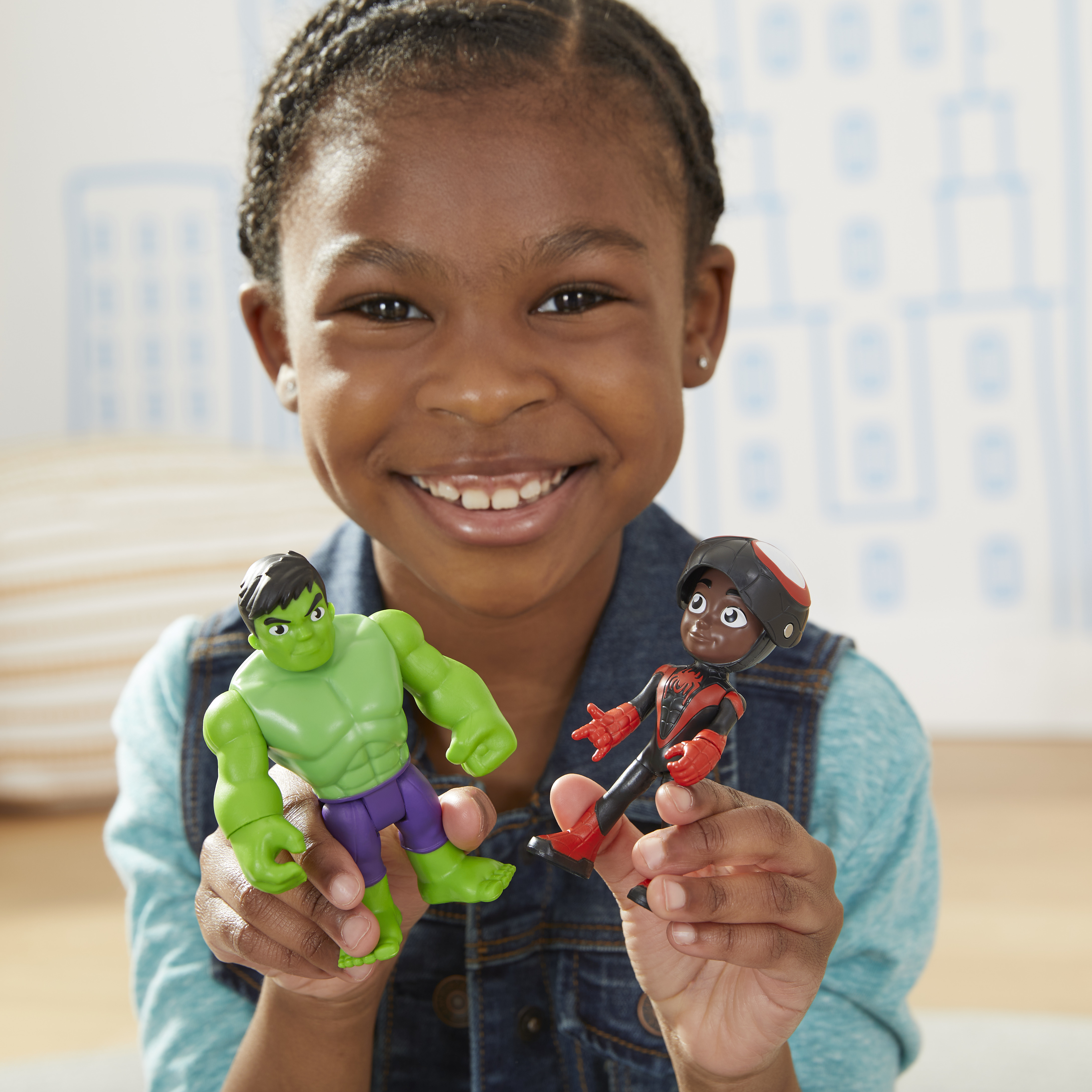 Una ragazza tiene due giocattoli: Hulk e Spider-Man. Sorride alla fotocamera. Sfondo: una città.
