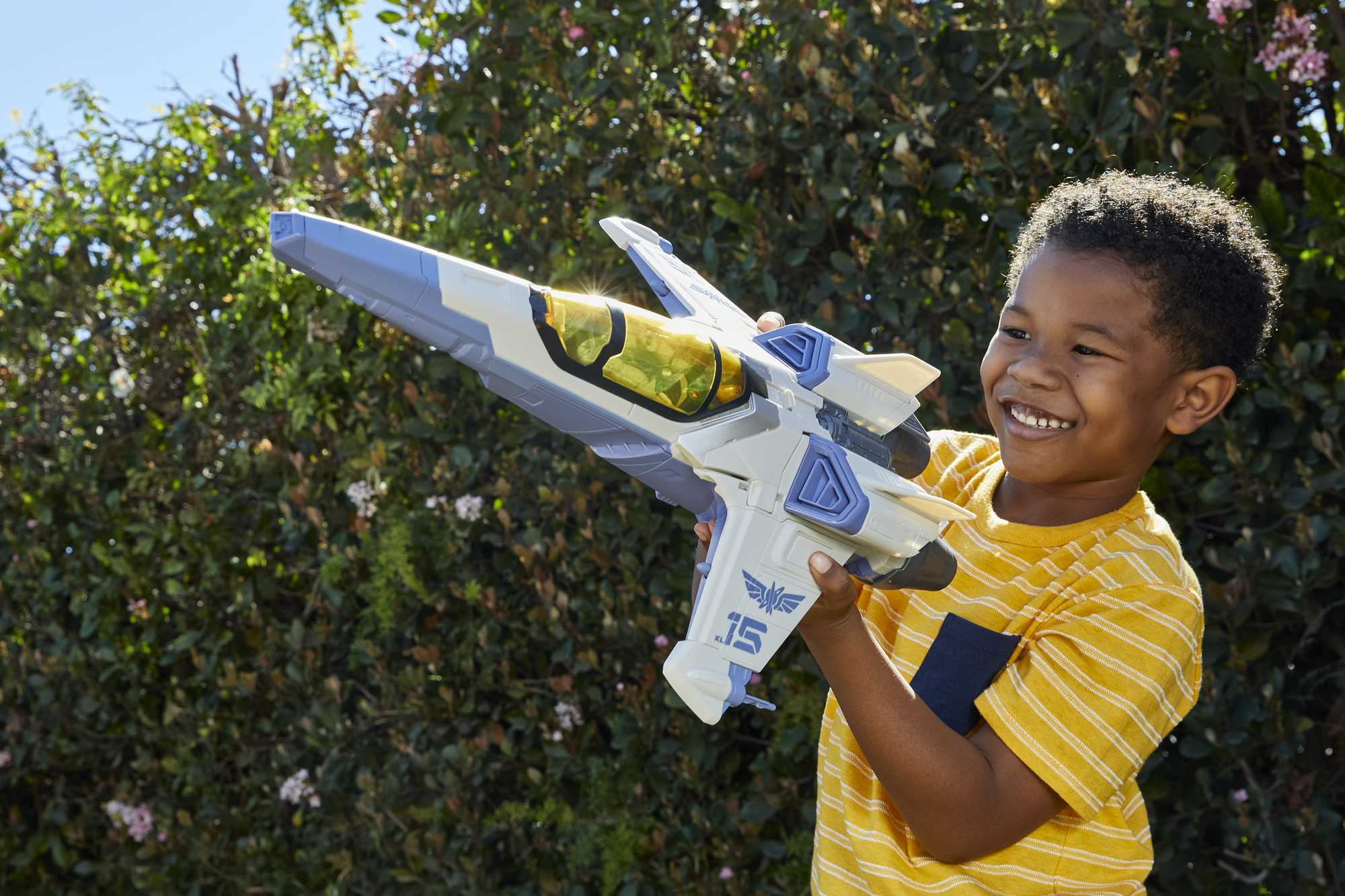 Un ragazzino che tiene un'astronave giocattolo, sorridente all'aperto con cespugli verdi.