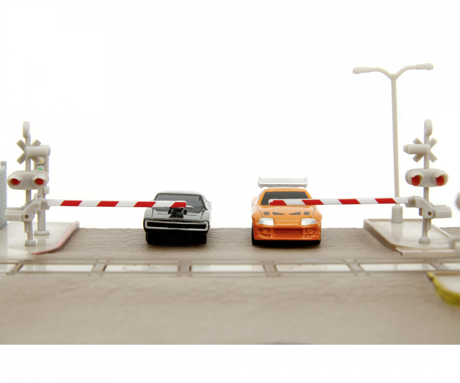 Zestaw zabawek z samochodami z dwoma samochodami, jednym czarnym, drugim pomarańczowym, na przejeździe kolejowym z szlabanami.