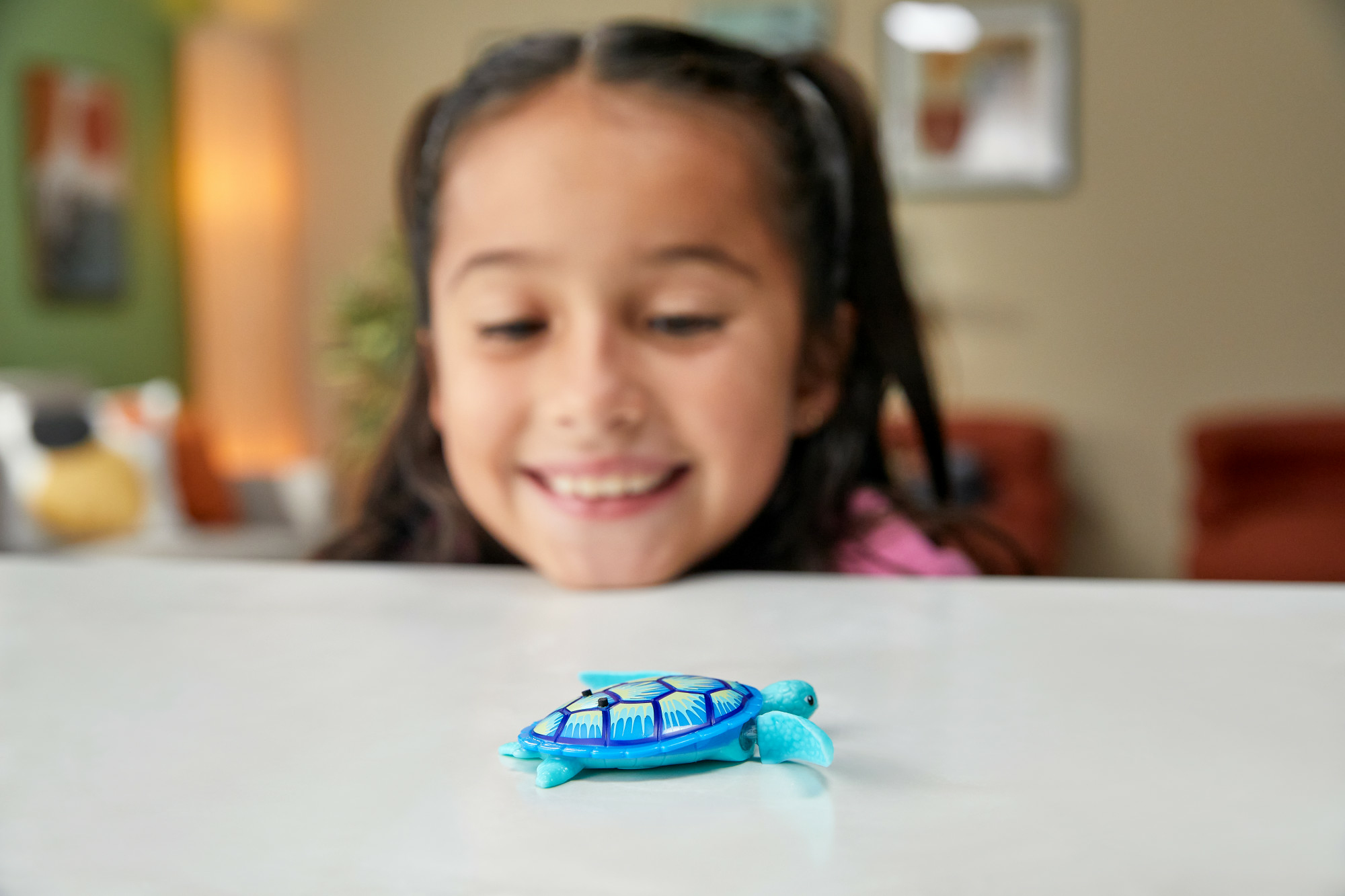 Una bambina sorride a una tartaruga giocattolo. La tartaruga è blu e verde, su una superficie bianca.