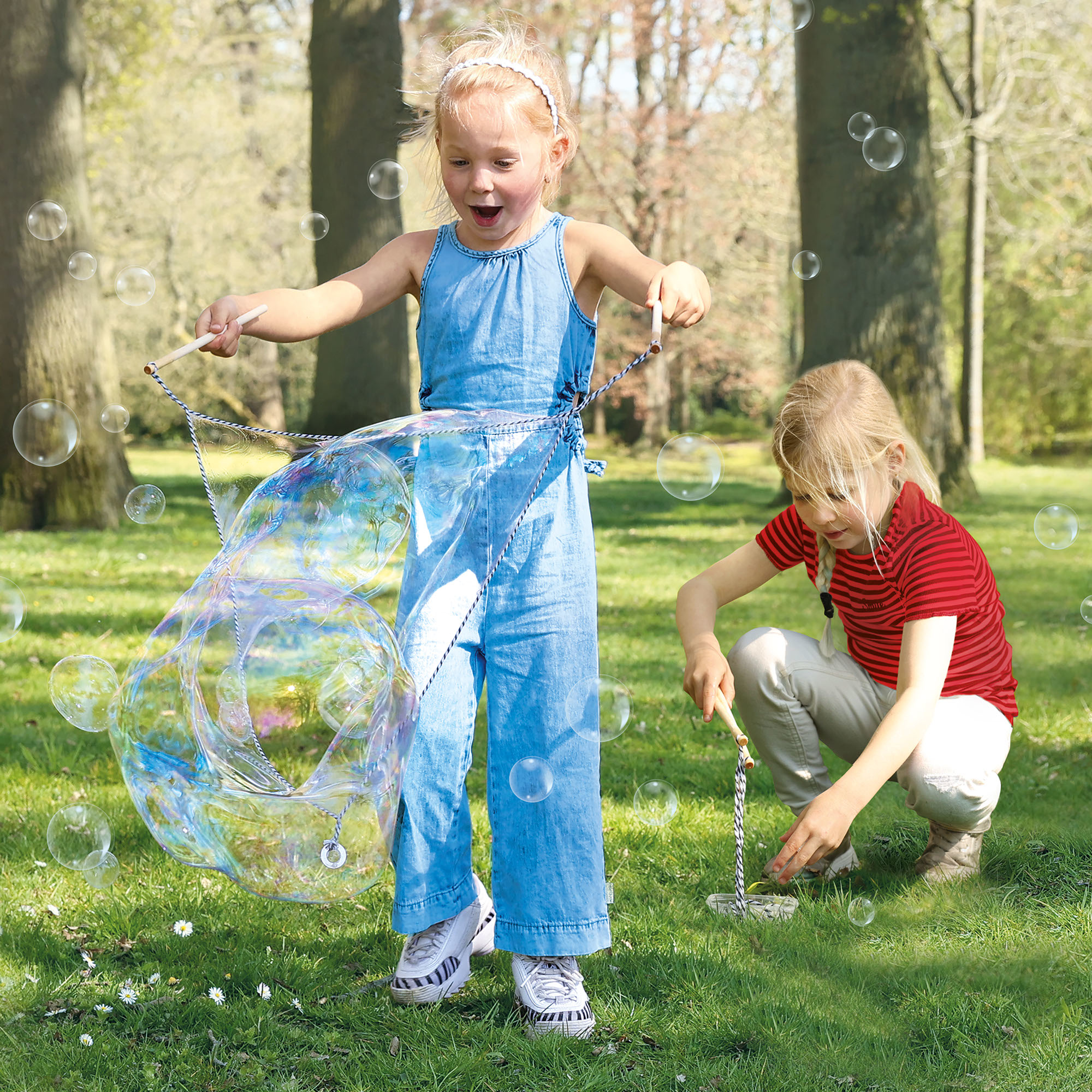 Due bambine che giocano con le bolle in un parco. Una tiene una bacchetta, l'altra immerge una bacchetta.