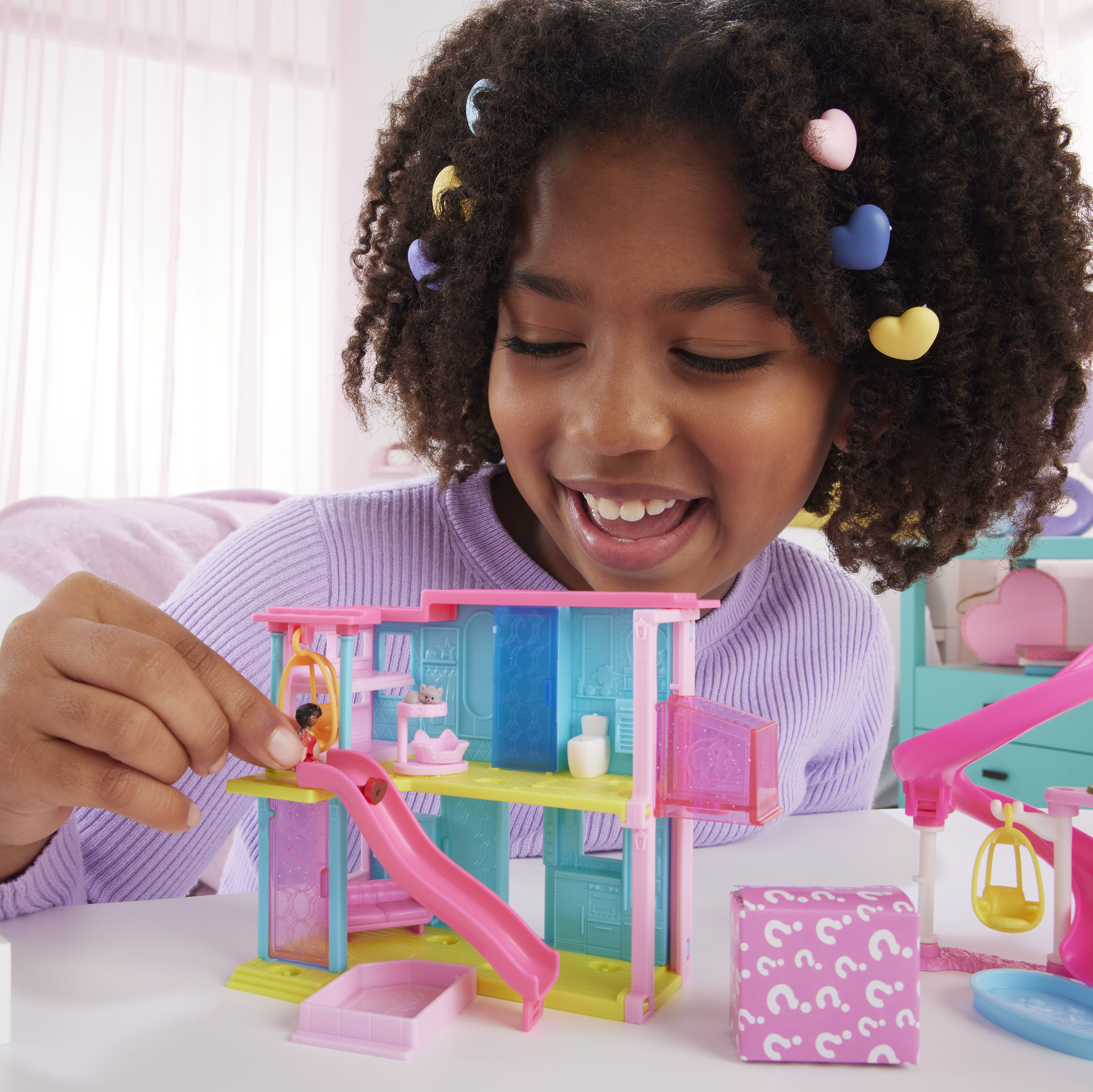 Dziewczynka bawi się zestawem Barbie. Widoczny jest domek ze zjeżdżalnią i łukiem tęczy. Dziewczynka się uśmiecha.