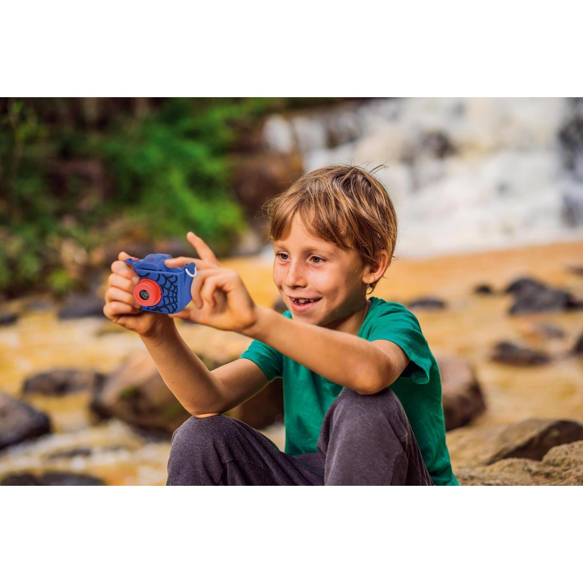 Chłopiec uśmiecha się, trzymając niebiesko-czerwony aparat na tle rozmytego wodospadu.