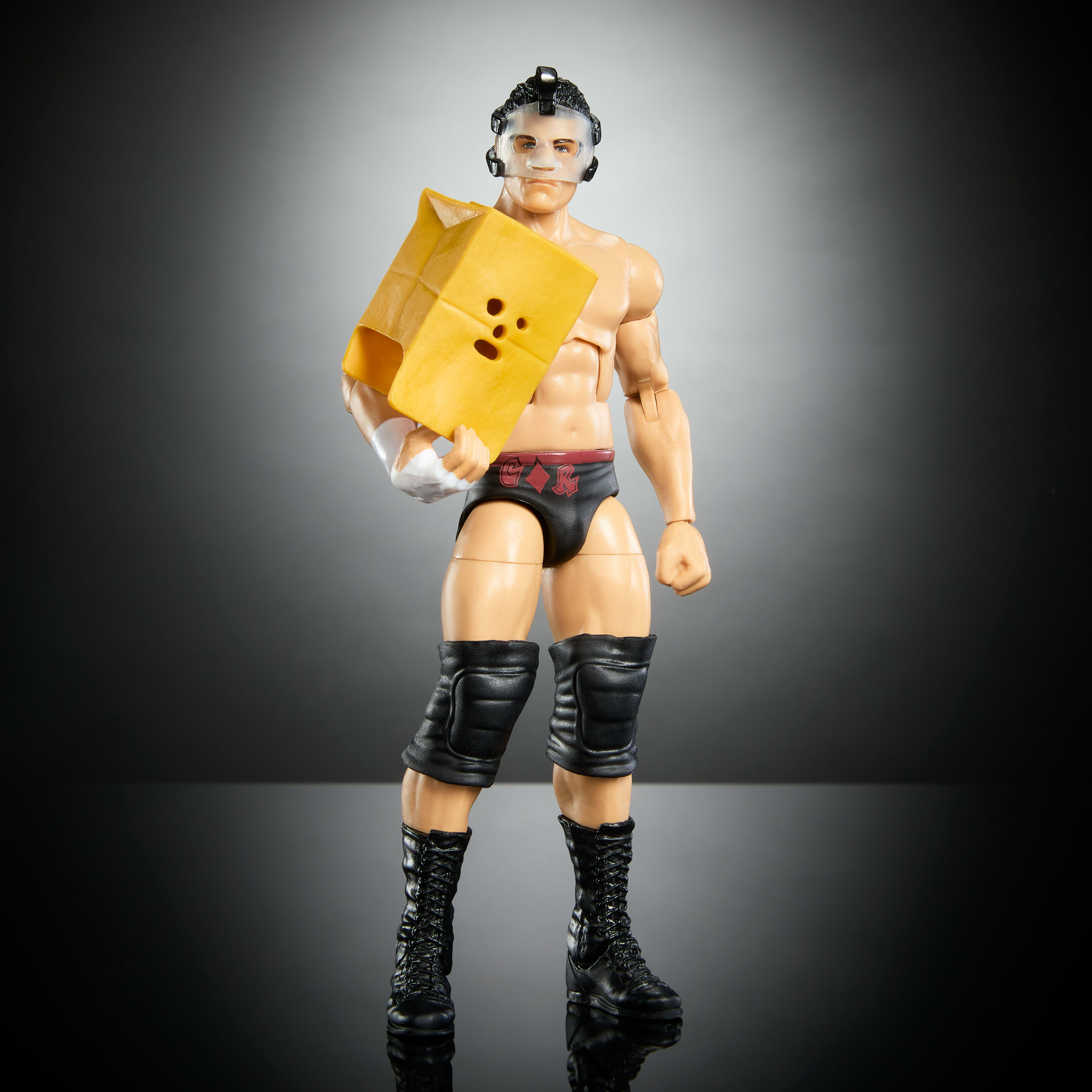 Figurina di wrestler con maschera, tiene una scatola gialla. Pantaloncini e stivali neri. Sfondo grigio.