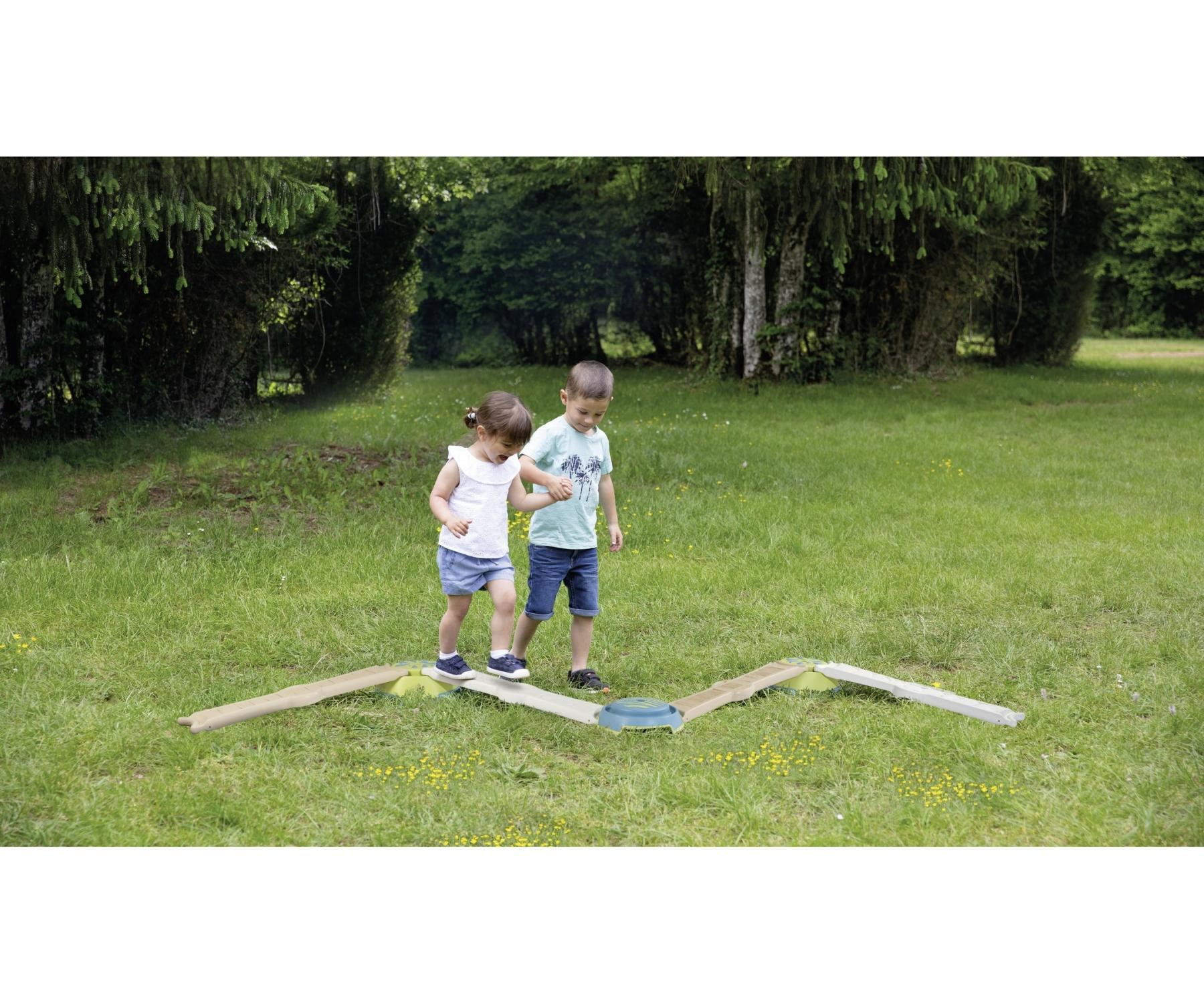 Two children walking on a balance beam in a grassy park. They are smiling and playing.