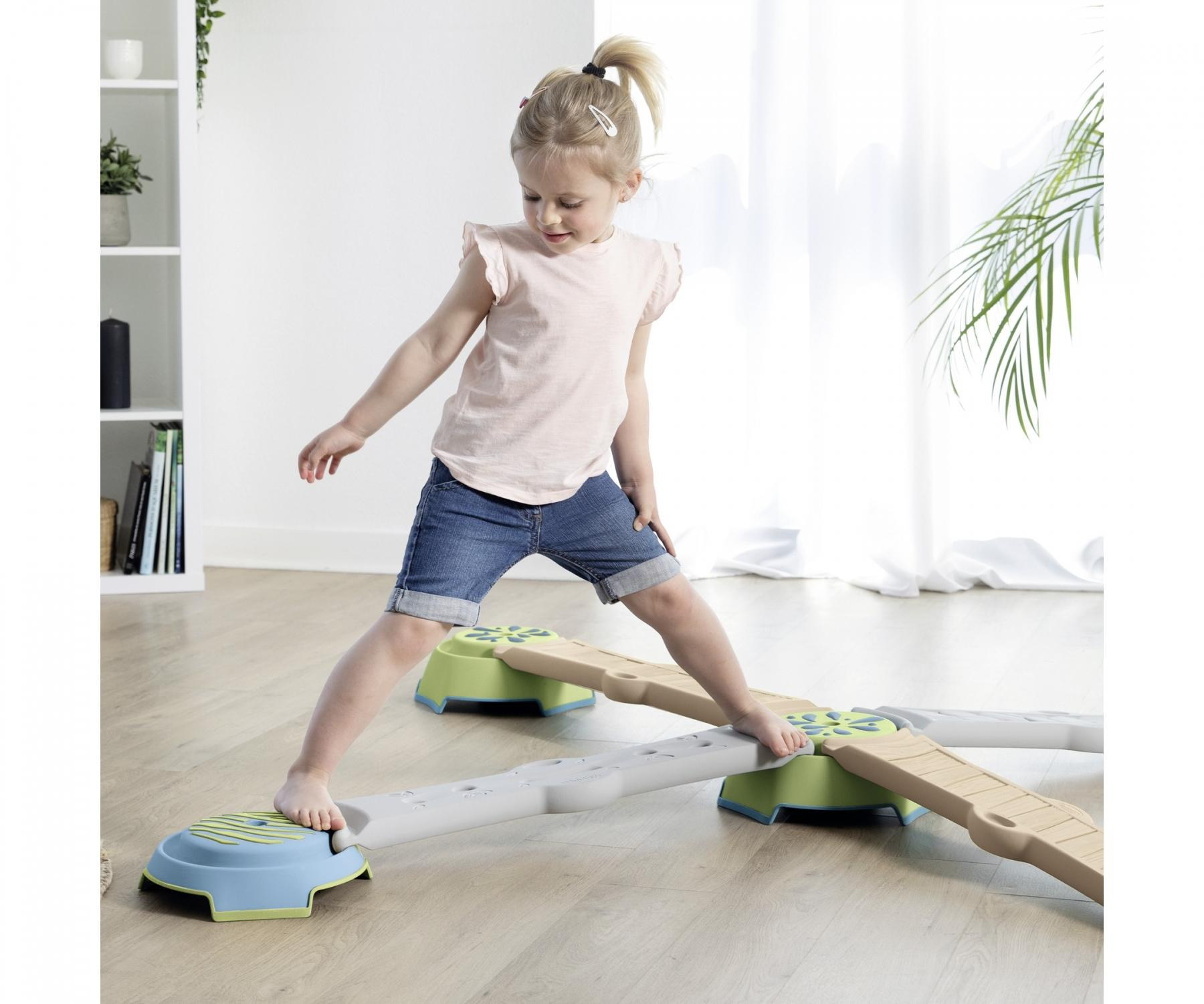 A young girl balances on a play structure with stepping stones. The structure is in a bright room.