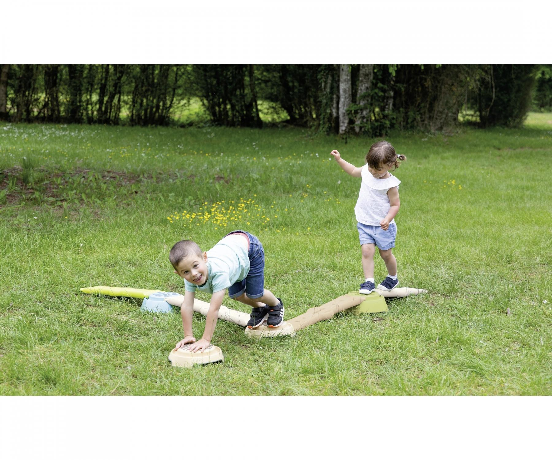 Two children play on a balance beam in a grassy area. One crawls, the other walks. Background is trees.