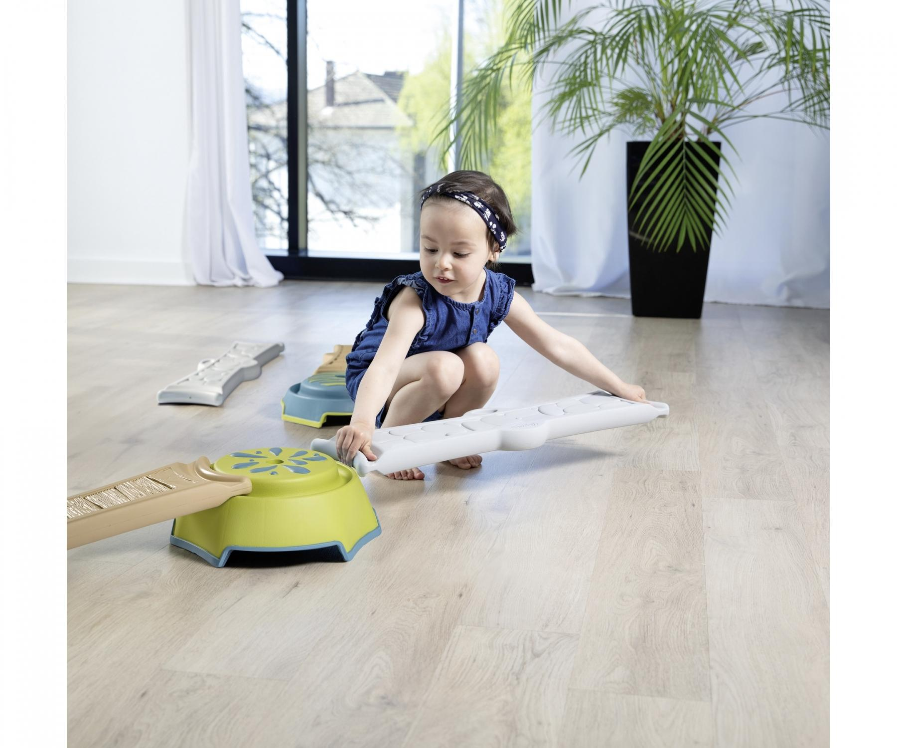 A young girl plays with colorful toy pieces on a wooden floor. A plant sits in the background.
