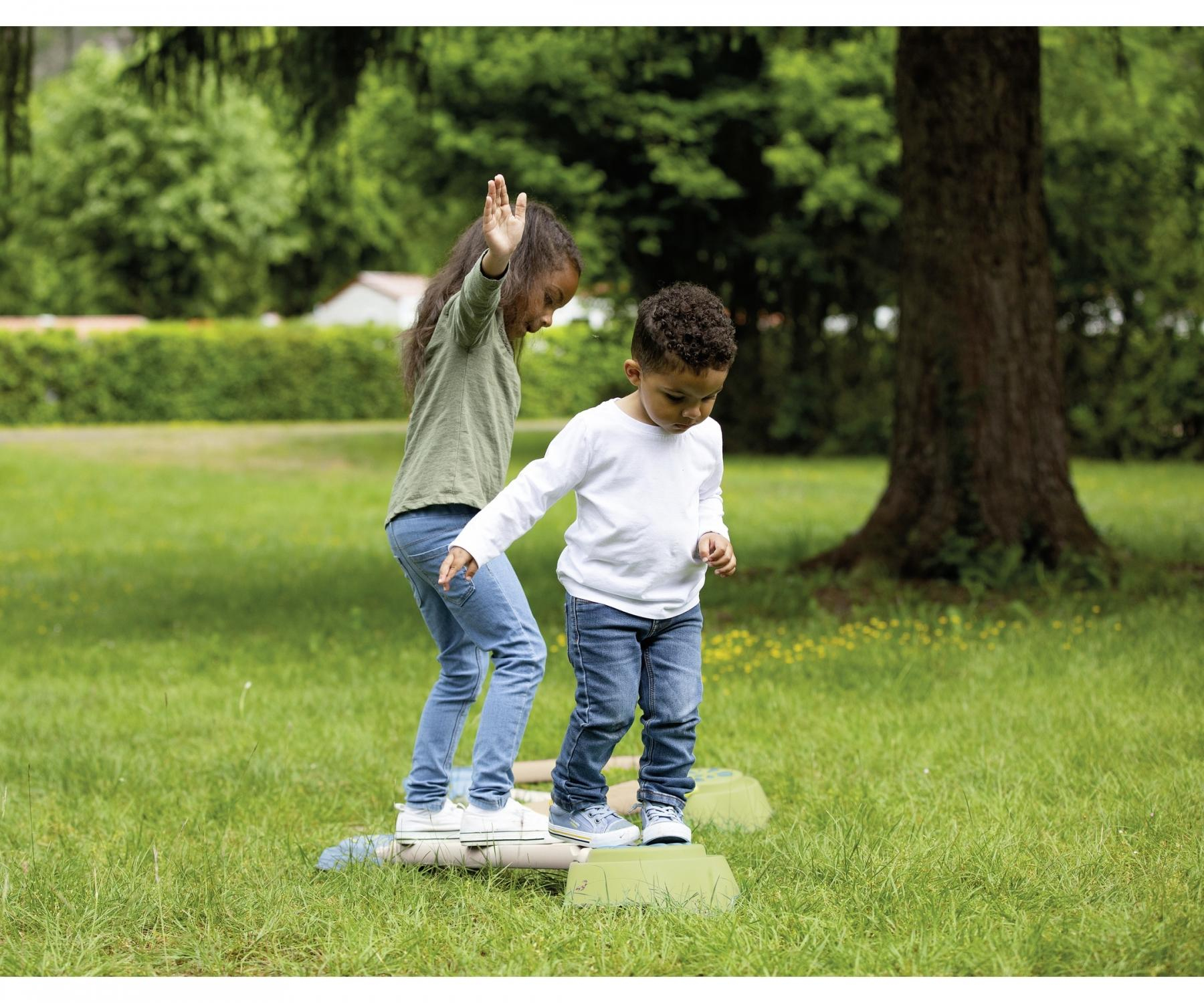 Two children playing on stepping stones in a green grassy area with a tree.
