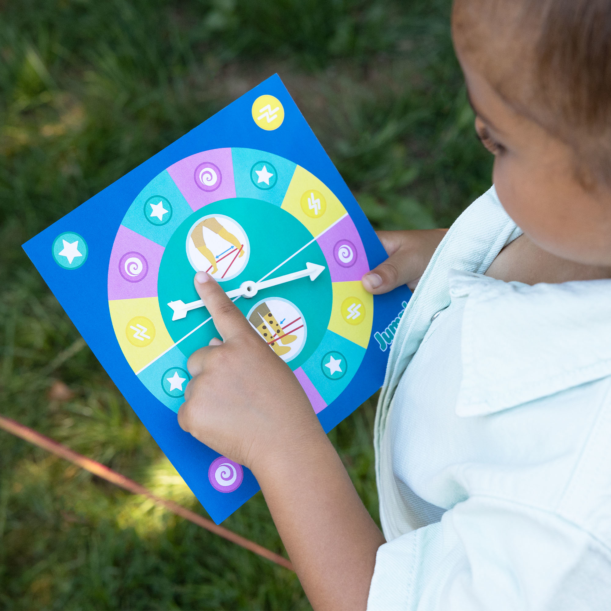 Un niño señala un juego de mesa con una ruleta. El juego es azul con secciones coloridas e ilustraciones. Hierba verde al fondo.