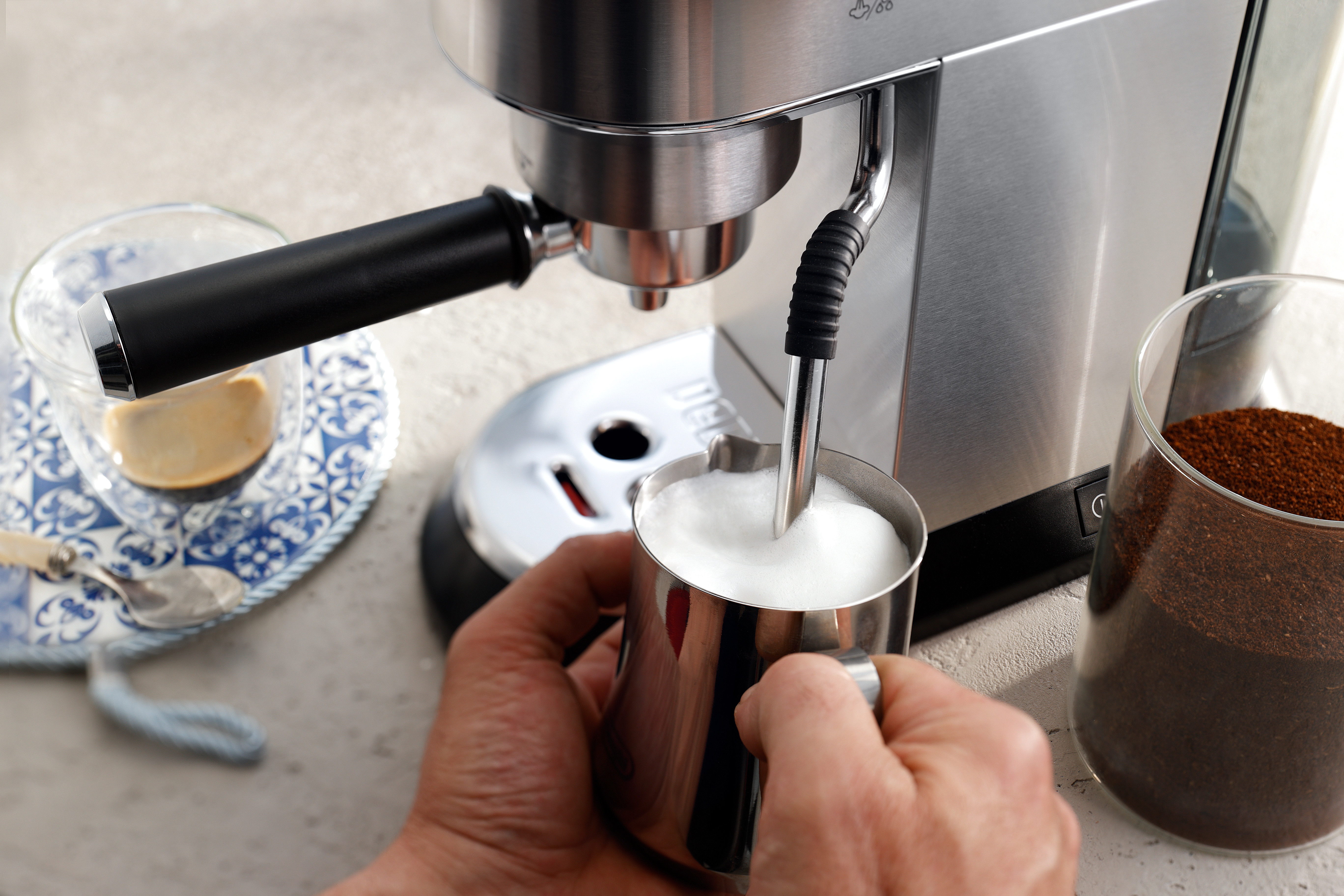 Eine Person schäumt Milch mit einer De'Longhi Espressomaschine auf. Es gibt ein Glas Kaffee und Kaffeepulver.