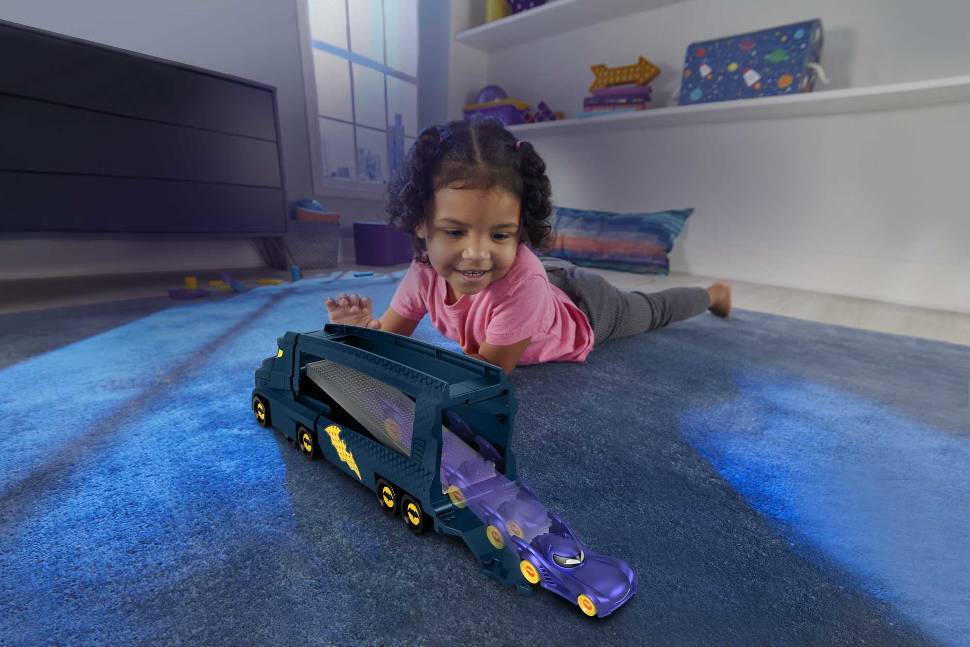 Dziewczynka bawi się niebieską zabawką ciężarówką Batmana. Fioletowy samochód wyjeżdża. Pokój ma okno i telewizor.