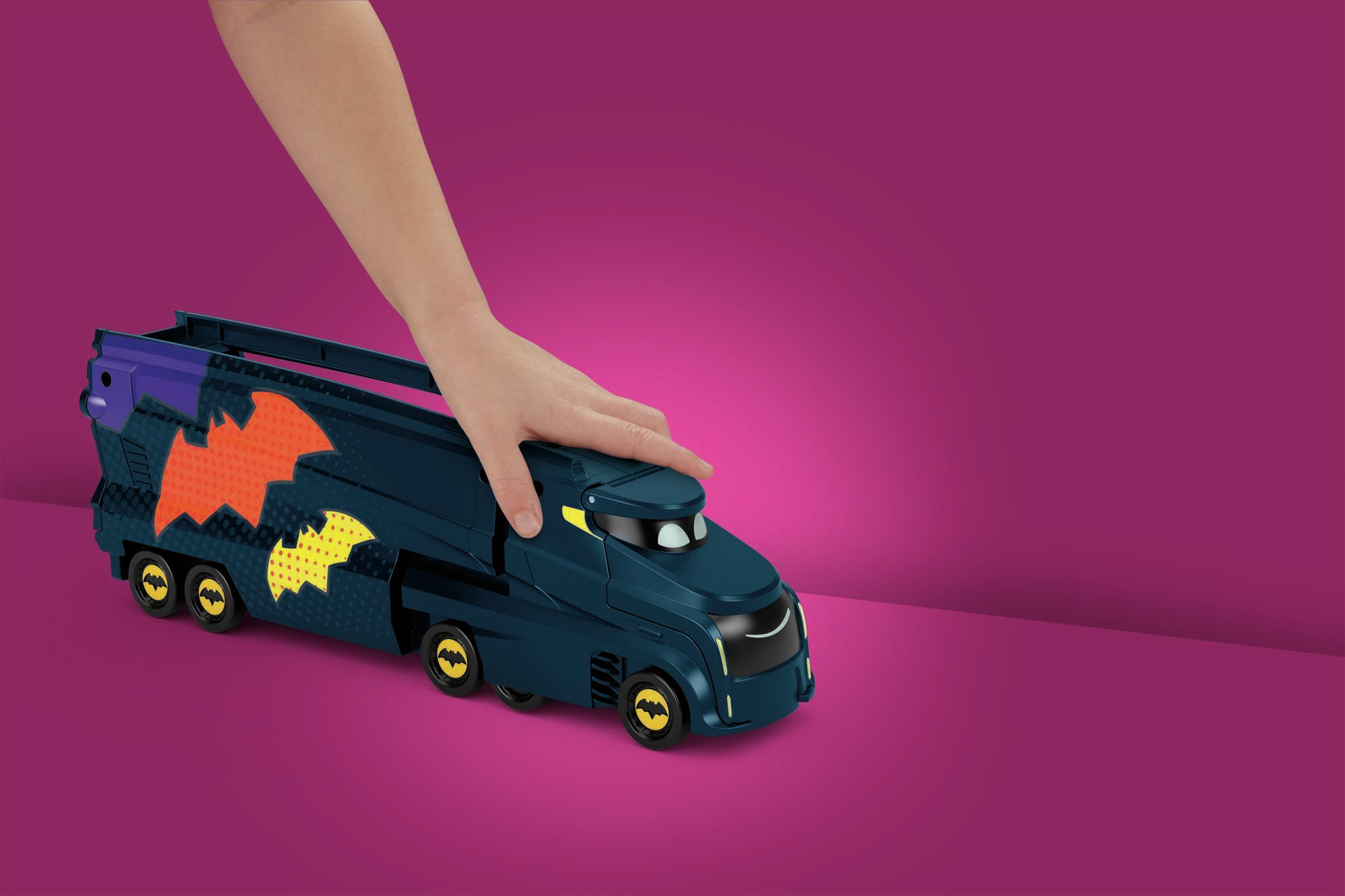 Ręka na niebieskiej zabawce ciężarówce Batmana. Ciężarówka stoi na różowej powierzchni. Logo Batmana na ciężarówce.