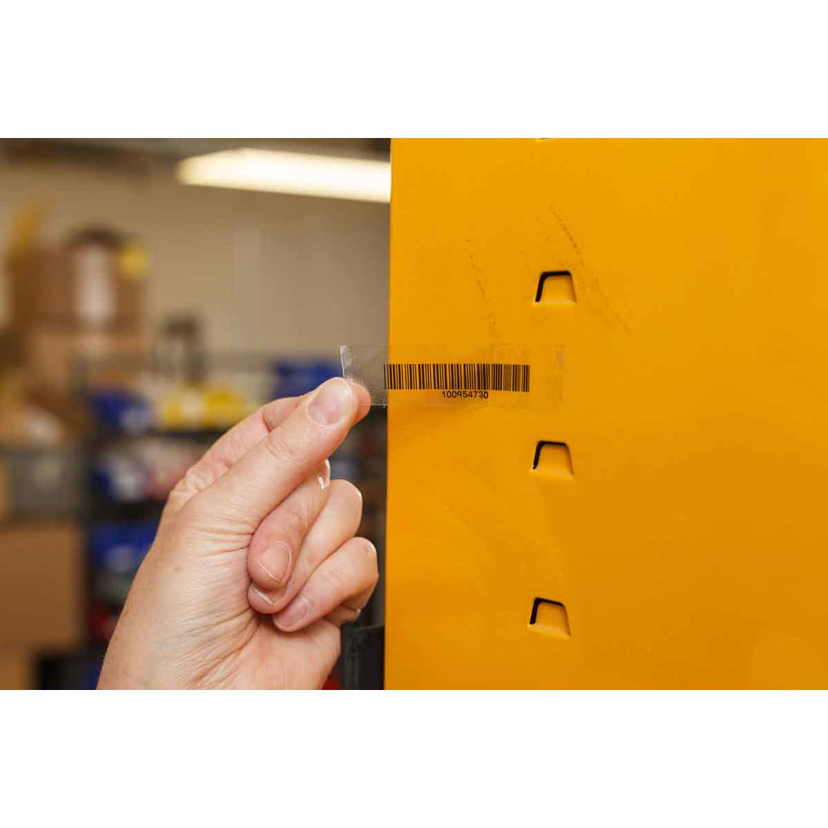 Dłoń trzyma przezroczystą etykietę z kodem kreskowym, w pobliżu żółtej szafki z otworami.
