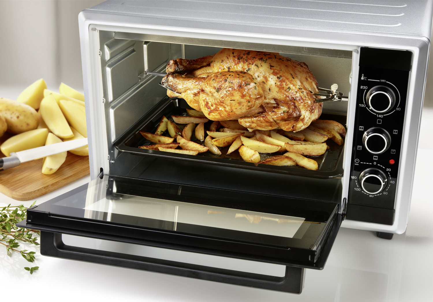 Piekarnik z pieczonym kurczakiem i frytkami. Ziemniaki i nóż na drewnianej desce. Drzwi otwarte.